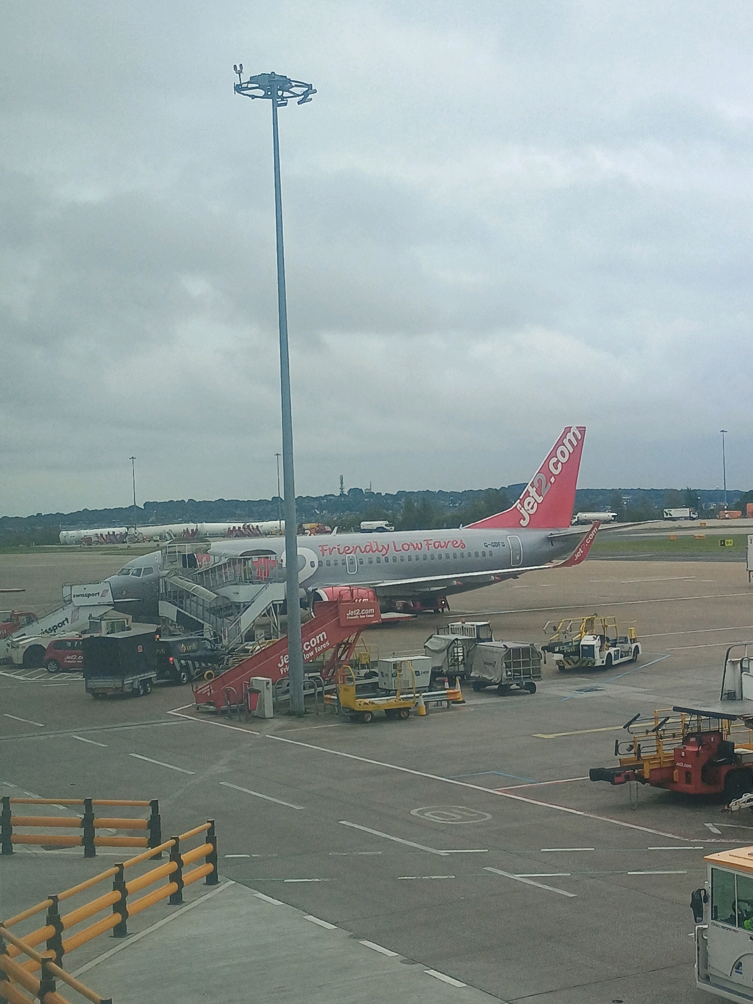 An aerial view of an airport tarmac on an overcast day, featuring a Jet2.com airplane parked at a gate. The plane's tail, adorned with the airline's signature red logo, is prominently visible. Several mobile stairs are connected to the aircraft, indicating passenger or crew activity. Various ground support vehicles, including baggage carts and a small tug, are positioned around the plane. In the background, other aircraft are visible parked on the tarmac, along with a long row of what appear to be airport lighting poles and a distant treeline under a cloudy sky. The foreground shows a section of the airport pavement marked with lines and bordered by yellow safety barriers.