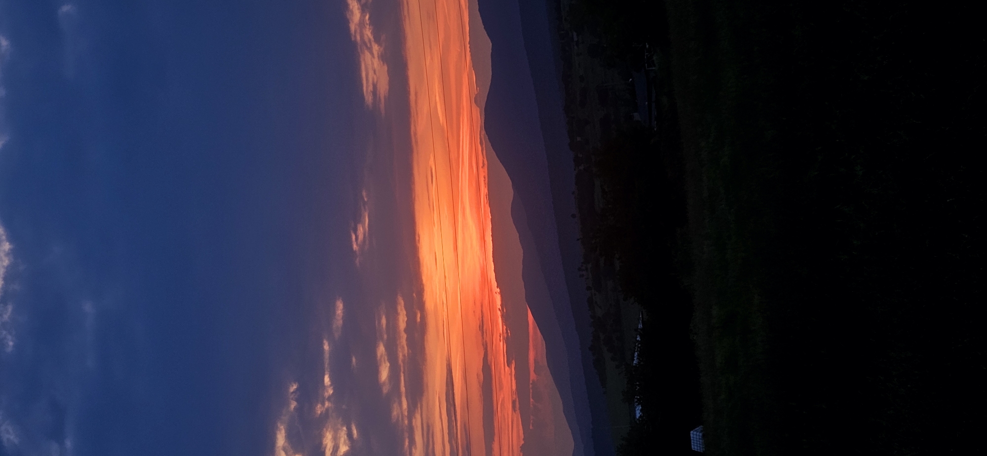The image depicts a sunset or sunrise over a mountainous, rural landscape in Momin Prohod, Bulgaria. The sky transitions from a dark indigo at the zenith to a vivid orange and red horizon. Sparse, dark cumulus and stratocumulus clouds are visible across the upper sky, with some illuminated by the warm light near the horizon. Multiple thin, dark horizontal lines, consistent with power or telephone wires, traverse the middle section of the vibrant orange sky.
Below the sky, several distinct layers of mountains are silhouetted in deep shades of blue and purple, increasing in lightness and blueness with distance due to atmospheric perspective. The immediate mid-ground reveals a dark, undulating terrain with indistinct vegetation and sparse, dark structures suggesting a rural settlement or scattered buildings. The foreground consists of dark, almost black, silhouetted vegetation, primarily grass and trees, casting deep shadows. On the far left of the foreground, a light-colored, rectangular object, possibly a solar panel or a tent-like structure with a corrugated surface, is visible. No human figures or discernible animal life are present. The scene is unlit by artificial sources, relying entirely on natural twilight illumination.