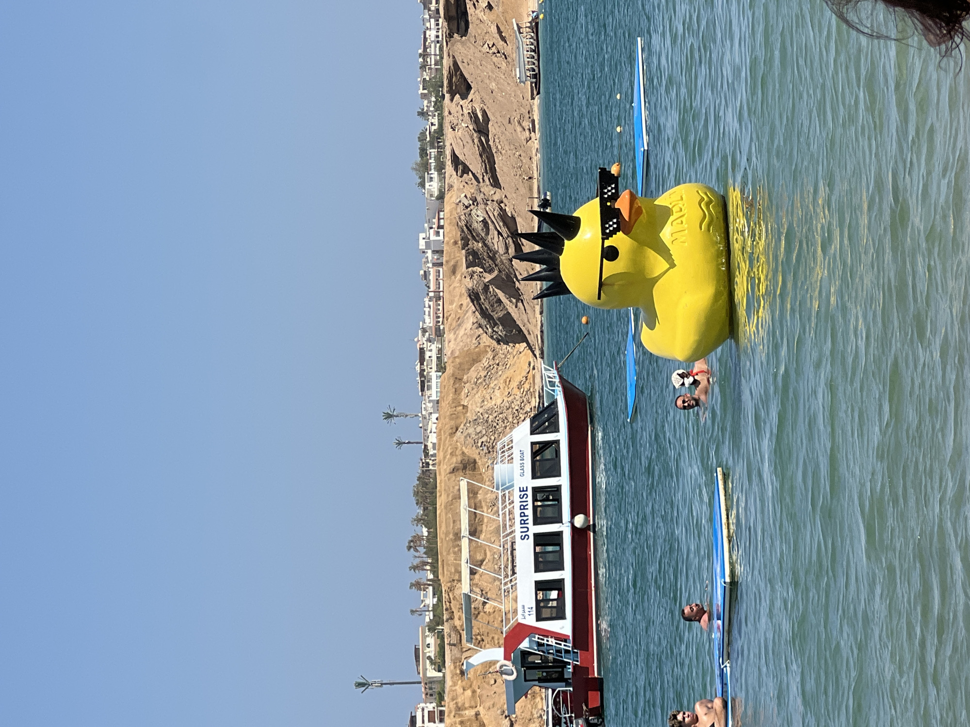 The image captures a sunny day at a beach or bay in Sharm El Sheikh, Egypt. In the foreground, a large, inflatable yellow duck with a mohawk and pixelated sunglasses floats on the turquoise water. To the left, a red and white boat, labeled "SURPRISE GLASS BOAT 114", is anchored. Several people are visible in the water, some on paddleboards, enjoying the day. In the background, a sandy, rocky coastline with buildings and palm trees lines the horizon under a clear blue sky.
