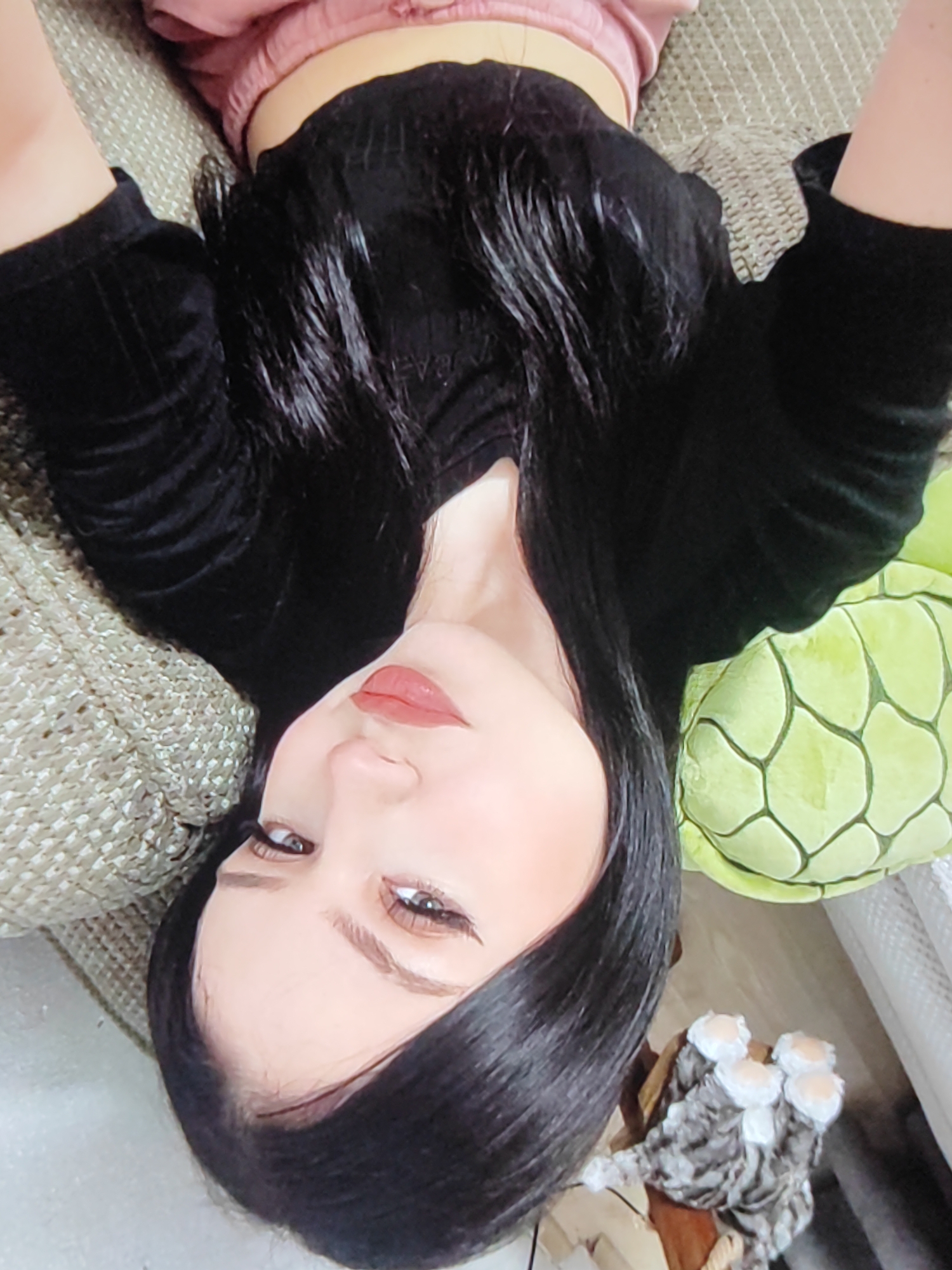 This is a media file featuring a person lying down on a textured surface, possibly a couch or bed. The person has long, dark hair and is wearing a black top and pink bottoms. Their face is turned towards the camera, and they appear to be looking directly at it.
The scene is indoors, with a light-colored textured surface as the background. To the right of the person, a green, patterned object, possibly a stuffed toy or cushion, is visible. Further to the right, a decorative item resembling a cluster of white and gray fluffy objects is also present.
The lighting suggests it is daytime. The person's expression is neutral, and there are no discernible emotions. No text is visible in the media file. The overall impression is a casual, personal snapshot. The location context is Fergana City, Uzbekistan.