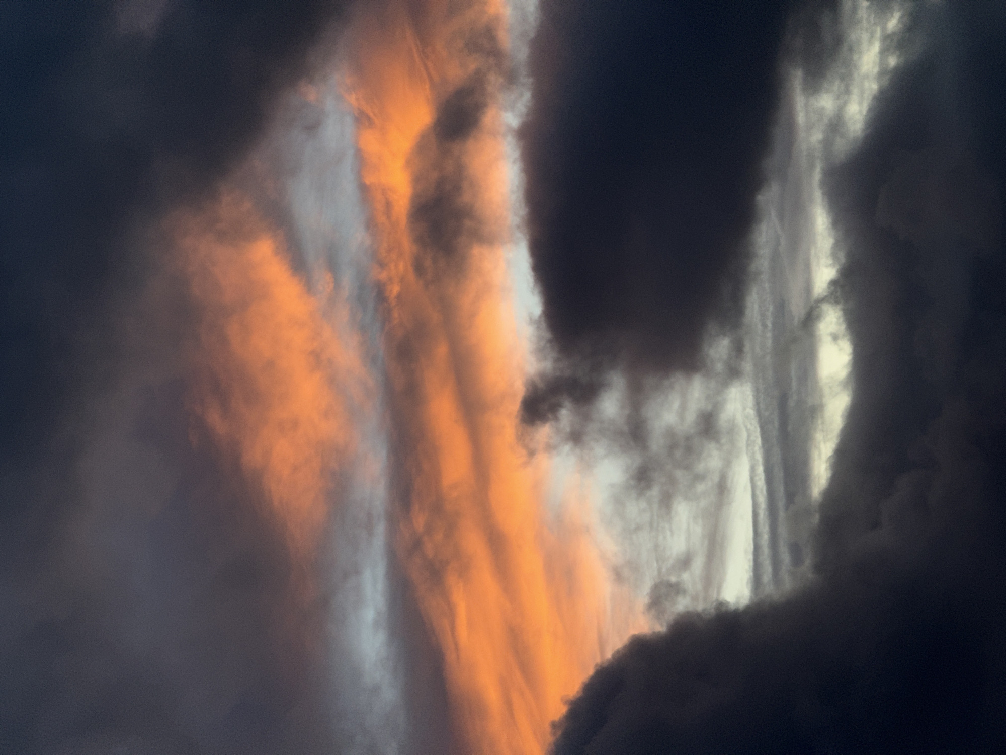 The image captures a dramatic and dynamic sky, likely during sunrise or sunset over Fécamp, France. The scene is dominated by an impressive array of clouds, showcasing a striking contrast between vivid light and profound darkness.
A prominent central band of clouds glows intensely with rich orange and amber hues, illuminated by the low-angled sun. These brightly lit clouds appear as swirling, elongated forms, stretching vertically across the frame, with some lighter white and pale gray sections interspersed within the vibrant orange. This luminous core is sharply bordered on both sides by heavy, dark gray to deep blue-black clouds, suggesting dense storm formations or deep shadows.
The textures of the clouds vary significantly; the orange-lit areas appear softer and more diffused, while the darker masses possess a more turbulent, dense, and opaque quality. The interplay of light and shadow creates a powerful sense of depth and movement in the sky. No specific objects, people, or discernible text are visible in the frame. The overall atmosphere is one of natural grandeur and dramatic beauty, hinting at powerful weather phenomena underscored by the fleeting beauty of dawn or dusk.