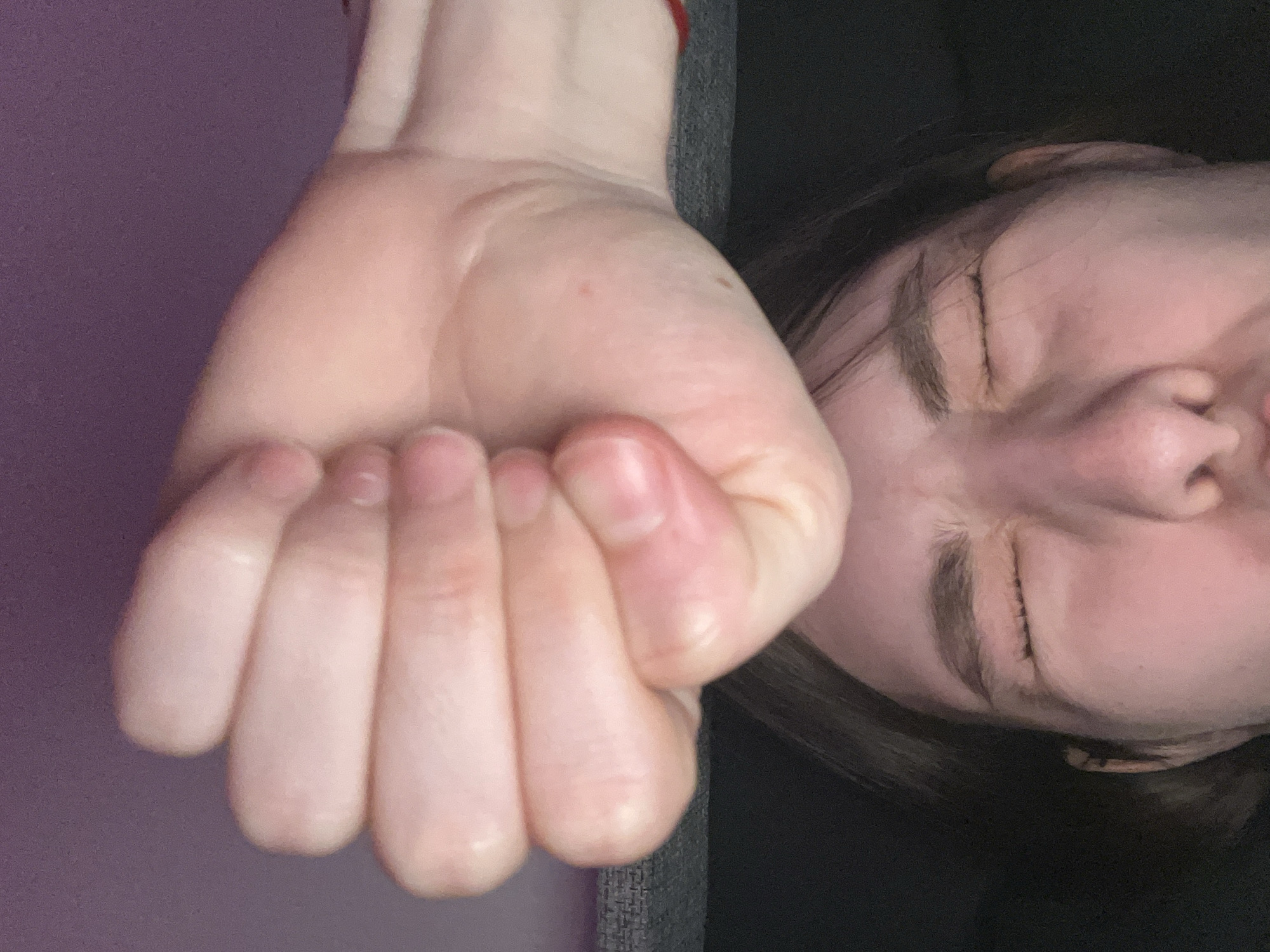 A person's face is visible in the upper right corner of the image, their eyes closed. Next to the face, in the left foreground, is a clenched hand. The background appears to be a combination of purple and grey colors. There is no information in the image to identify the location.