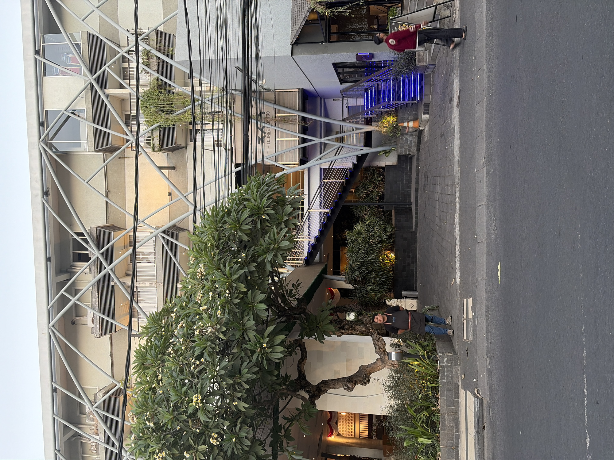 The image depicts a street scene in Kuta, Indonesia, featuring a modern building with a distinctive geometric metal facade and multiple balconies. A large tree with white flowers is prominently positioned in the foreground to the left.
There are two individuals visible. On the left, a person wearing an apron and jeans stands near the tree. On the right, another person in a red jacket and dark pants walks away from the viewer.
The building shows several levels with balconies, some adorned with hanging plants. A brightly lit blue staircase leads up to an entrance. Overhead, a dense network of electrical wires crisscrosses the frame. The street in the foreground is paved with asphalt, and the sidewalk area is constructed of interlocking pavers. The environment appears to be a commercial or hospitality district, with indications of businesses like "ZA LOUNGE & BISTRO" visible on signage. The overall lighting suggests it is either early morning or late afternoon.
