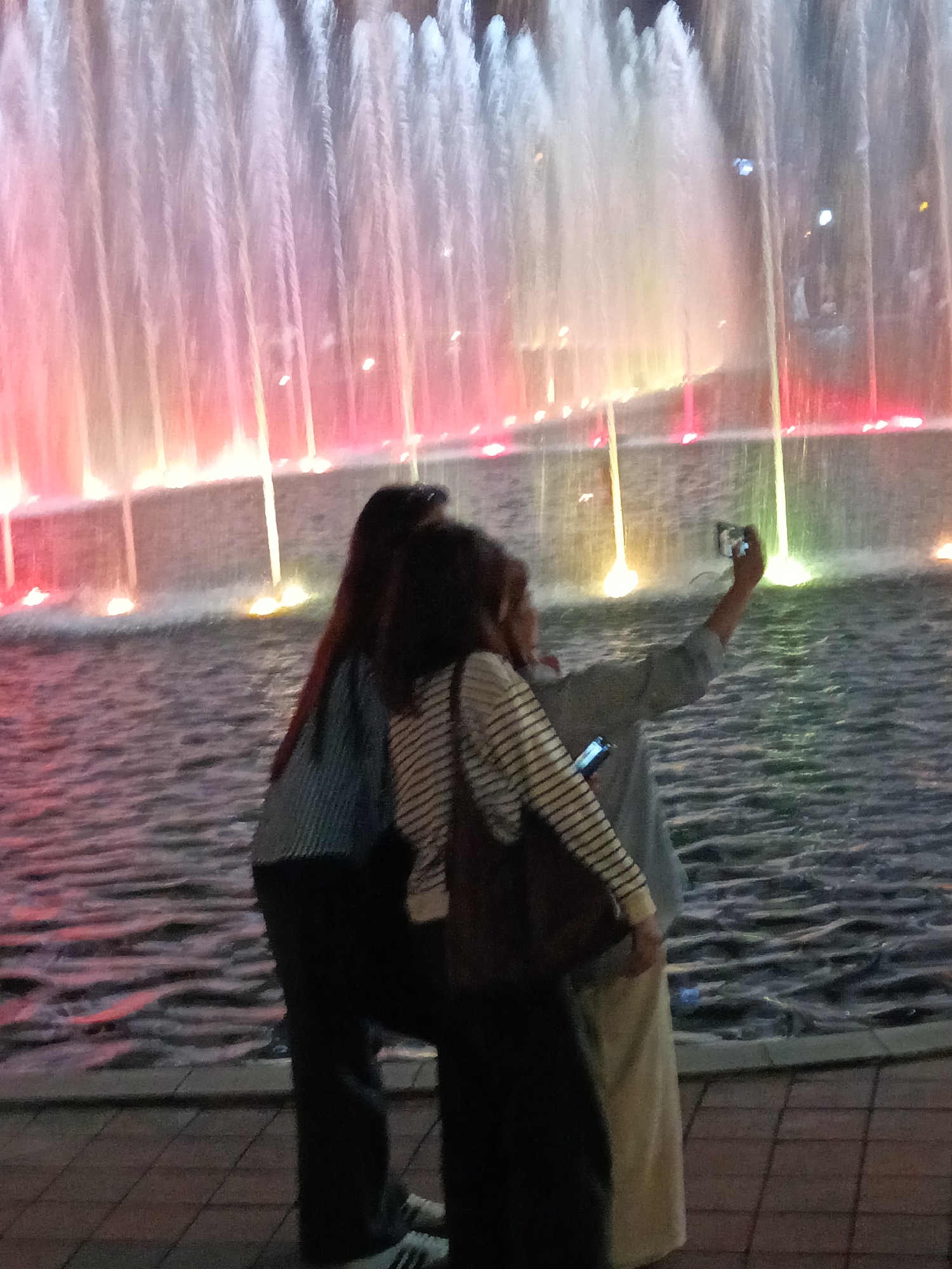 Two individuals are taking a selfie with a brightly illuminated fountain in the background. The fountain features cascading water illuminated with pink, white, and yellow lights. The individuals are positioned in the foreground, with one holding a smartphone to capture the image. The environment suggests an outdoor public space, likely a park or promenade, with paving stones visible in the lower portion of the frame and water from the fountain extending into the foreground. The overall impression is that of a nighttime scene. This could be the Kuala Lumpur City Centre (KLCC) Park, known for its Symphony Lake with a spectacular fountain show.