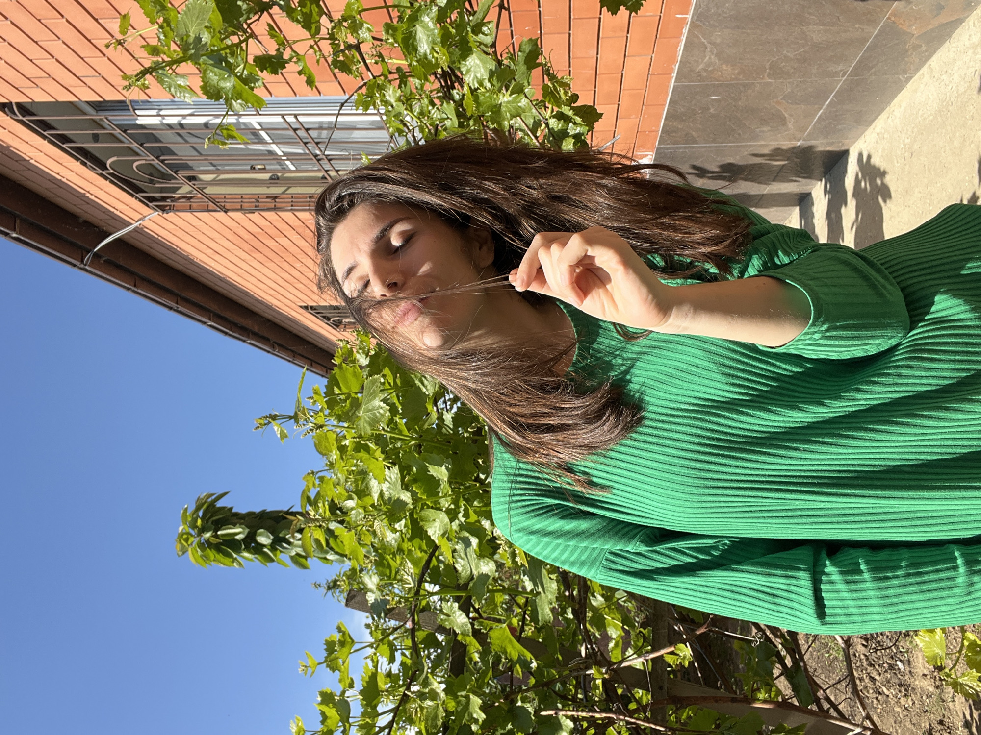 The image depicts a woman in a green ribbed top, standing outdoors. She has long brown hair and is holding a strand of it near her face with her right hand. Her eyes are closed. In the background, there is a brick building with a window that has a metal grate. Green vines with leaves are visible, obscuring some parts of the background. Above the building and vines, there is a clear blue sky. The image appears to be taken on a sunny day, given the shadows and brightness. Based on visual references and available information, the location is Kaspiysk, Russia.