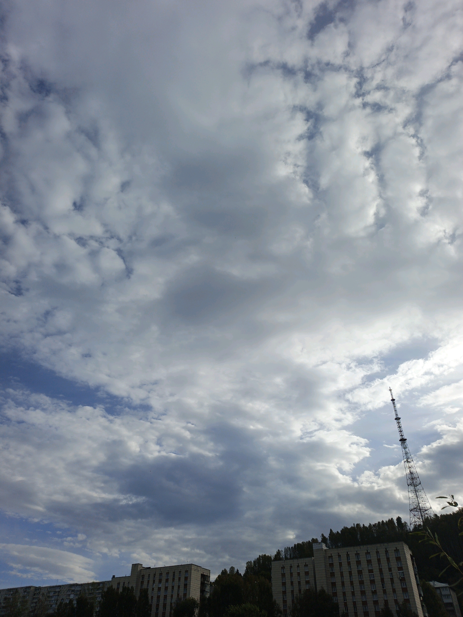 The image captures a cloudy sky above a cityscape in Zlatoust, Russia. In the foreground, a multi-story building with many windows is visible, partially obscured by trees. To the right, a tall telecommunications tower stands prominently against the sky, with the forested slopes of a hill behind it. The clouds are a mix of white and gray, suggesting a partly cloudy or overcast day. The lighting indicates it is daytime.