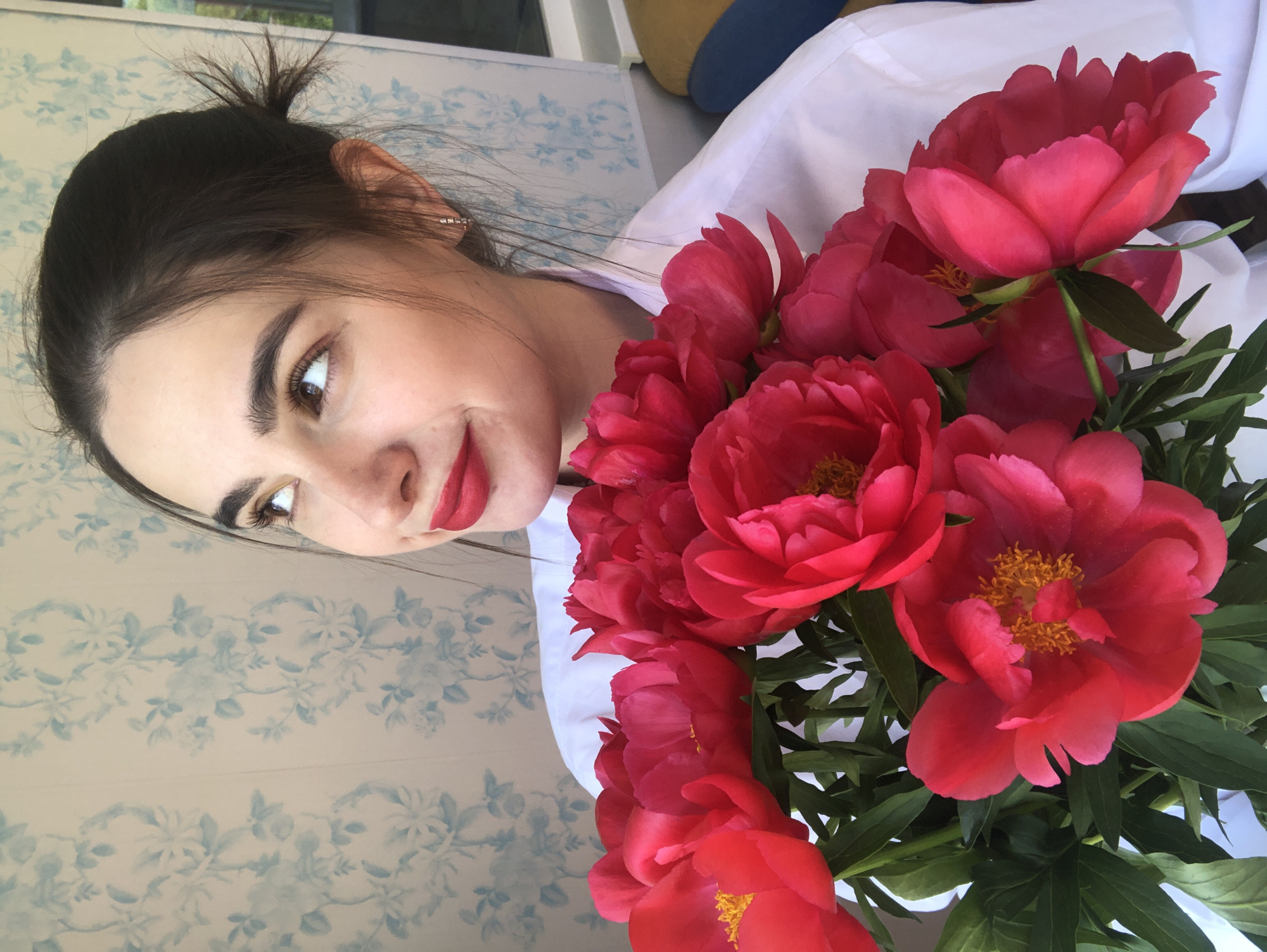 A young woman with dark hair pulled up into a messy bun and wearing a white collared shirt is holding a bouquet of bright pink peonies. She is looking upwards and to the right with a slight smile. Her lips are painted with red lipstick. The background features a light-colored wallpaper with a blue floral pattern. The image appears to have been taken indoors, possibly in Noviy, Russia, given the context provided. The lighting suggests natural light coming from the left side of the frame.