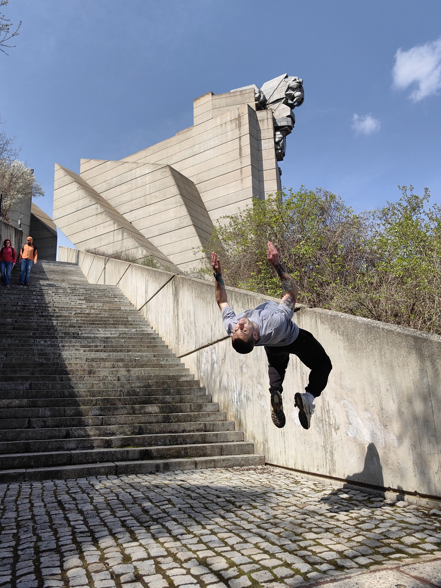 A man is mid-air, performing a backflip, in front of the Monument to 1300 Years of Bulgaria in Shumen, Bulgaria. The monument's concrete architecture is visible in the background, including the large stone sculptures at the top. Two individuals are walking up the concrete stairs leading to the monument. The ground is paved with cobblestones, and a concrete wall is to the right. Trees are visible in the background.