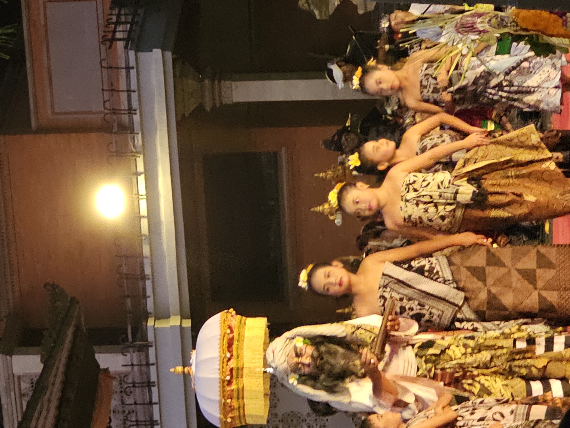 This media file captures a vibrant performance, likely a traditional Balinese dance, taking place outdoors at night, possibly in Denpasar, Indonesia. The scene is illuminated by a bright, warm light, casting a soft glow on the elaborately dressed performers and intricate surroundings.
The primary subjects are several young women, adorned in traditional costumes featuring patterned fabrics and floral headpieces. They appear to be dancers, their poses suggesting graceful movement and storytelling. One performer, positioned in the foreground, is more prominently featured, with a white and gold conical headdress, possibly a ceremonial object, placed beside her. Another figure, seemingly an older man with dark, long hair and a white head covering, is partially visible, his face expressing a serious or perhaps theatrical demeanor. The performers' attire is rich with detail, incorporating various textiles and ornamentation.
The setting seems to be an outdoor stage or courtyard with architectural elements that hint at a traditional Indonesian structure. A warm, bright light source, possibly a large lamp, is visible on the left side of the frame, illuminating a portion of the backdrop which appears to be a building with textured walls and decorative railings. The overall atmosphere is one of cultural celebration and artistic expression, with the night setting adding to the mystique and drama of the performance. The expressions on the faces of the performers, particularly the young women, convey a sense of focus and poise.