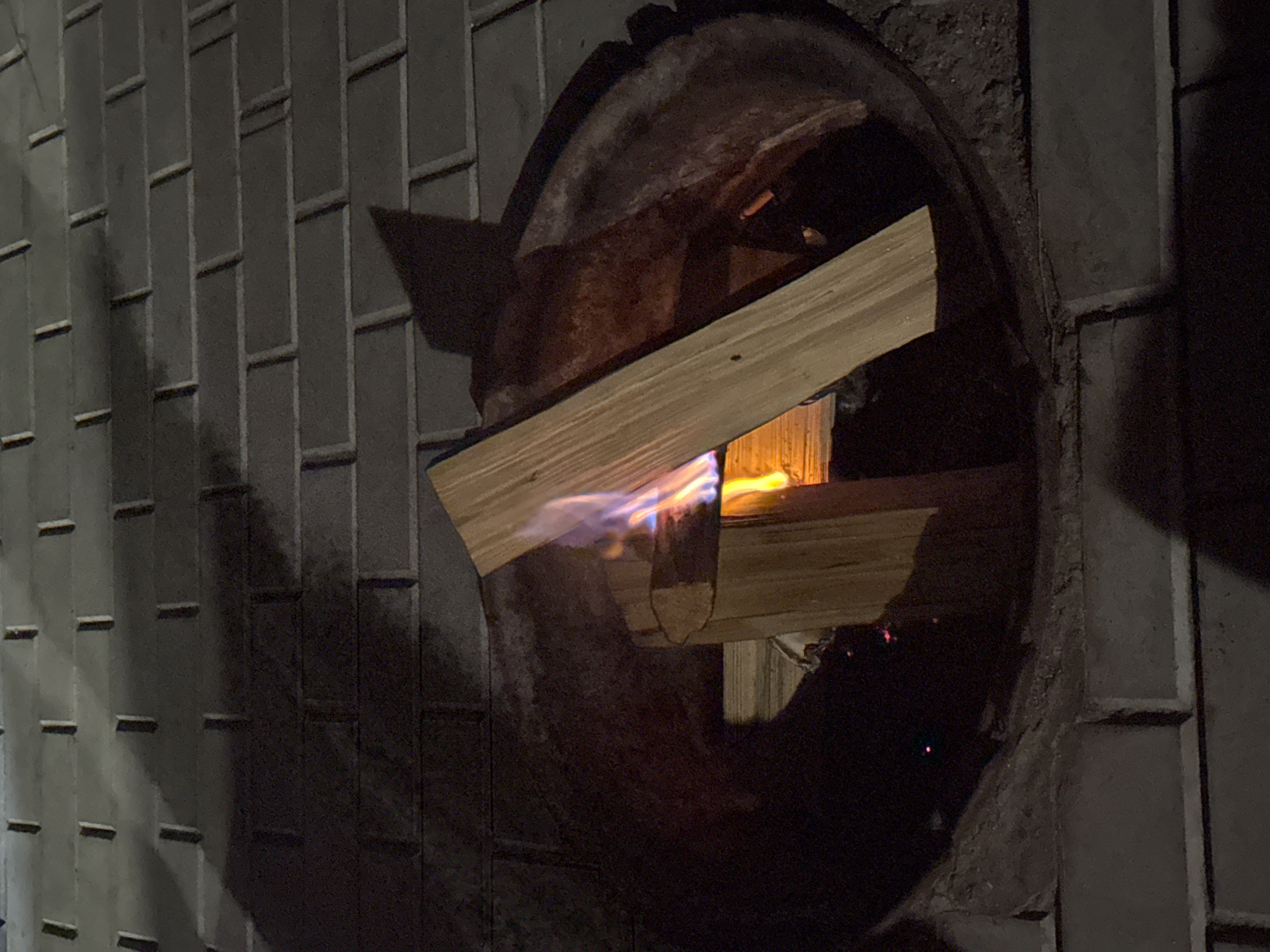 A medium shot captures a fire pit on a brick patio at night. Two pieces of firewood, arranged in an L-shape, are burning in the center of the metal fire pit. Flames, predominantly yellow and blue, lick at the wood. The surrounding brickwork is visible, with shadows cast from an unseen light source. The scene is set in Kurkut, Russia.
