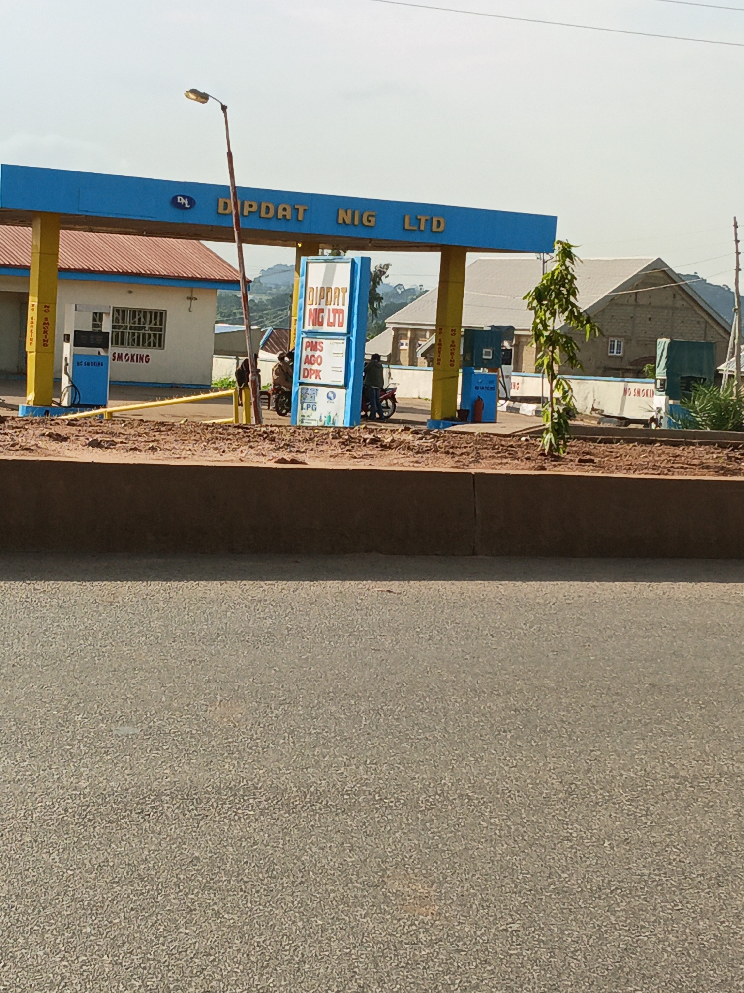 A DIPDAT NIG LTD gas station is visible in Bukuru, Nigeria. The station has a blue canopy with "DIPDAT NIG LTD" written on it in yellow letters. There are gas pumps under the canopy, and a white building with a red roof is behind the pumps. A sign lists prices for PMS, AGO, and DPk. Several people are present at the station, some near motorcycles. The ground is paved, and there is a concrete barrier in the foreground. The overall scene suggests a typical operational gas station in a Nigerian city.