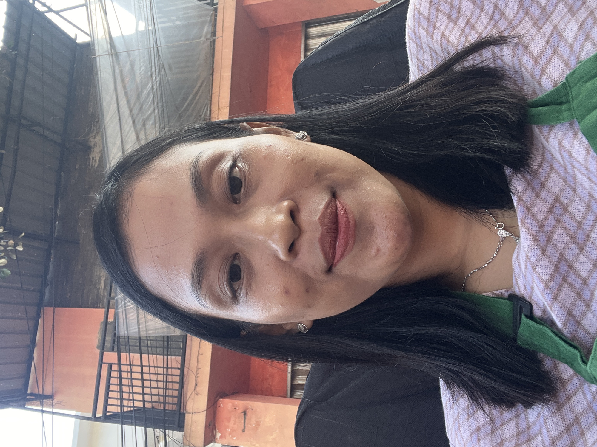 A medium-shot, outdoor selfie is captured from a frontal perspective, featuring a woman sitting in a chair. She has long, dark hair that extends to her shoulders. Her skin has a warm tone, and she is wearing subtle makeup, with a matte brown lipstick. She has on a light-colored, patterned sweater and a green strap across her chest. She is also wearing a silver necklace with a small pendant and small, sparkly earrings. The background shows an orange building with a black metal railing and a corrugated roof overhead. The scene is illuminated by natural daylight.