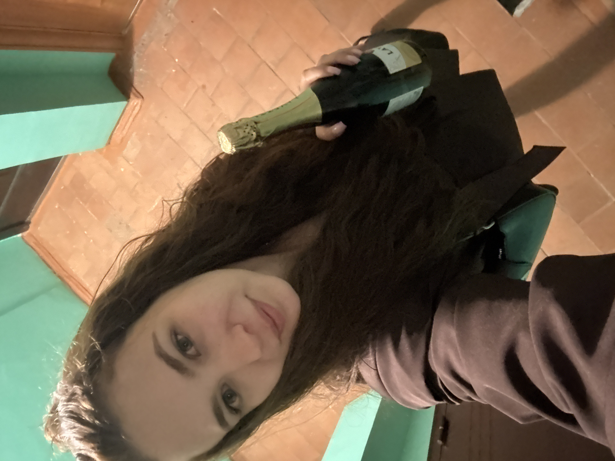 A young woman with long, brown hair holds a bottle of champagne. She is indoors, near a wall painted in a light green color. The floor is covered with orange-brown tiles. A black bag sits on a stool. The image was likely taken in Chaikovsky, Russia.