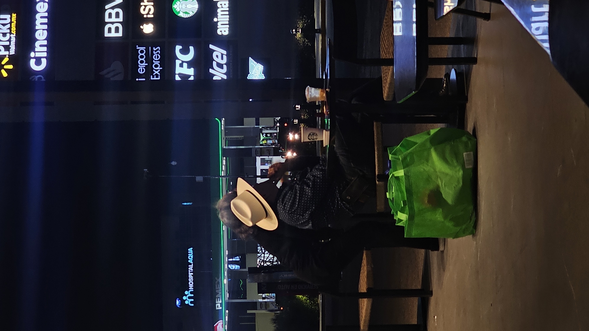 Here is a detailed description of the image:
**General Description**
The photo appears to be taken at night in an urban outdoor setting. The dominant features include illuminated store signs and a person sitting at a table with a shopping bag. The location is Heroica Puebla de Zaragoza, Mexico, as evidenced by the store signs visible in the photo.
**Specific Details**
* **Environment:** The scene is outdoors, likely a public area near a shopping center or strip mall. The time of day is night, indicated by the darkness and illuminated signs.
* **People:** A person is sitting at a table. This individual is wearing a light-colored hat, a dark jacket over a shirt with a patterned design.
* **Objects:**
* A bright green shopping bag is on the ground near the table.
* Several disposable coffee cups are on the table.
* Store signs are visible in the background. These include "PICKU," "Cinemex," "BBV," "Liverpool Express," "KFC," "Icel," "animal," "PEMEX," and "HOSPITAL AQUA."
* **Actions/Interactions:** The person is seated at the table, seemingly spending time there. There is a second person near the subject with the tan hat.
**Location and Context**
The store signs indicate the location is Heroica Puebla de Zaragoza, Mexico. The presence of multiple stores suggests a commercial area.