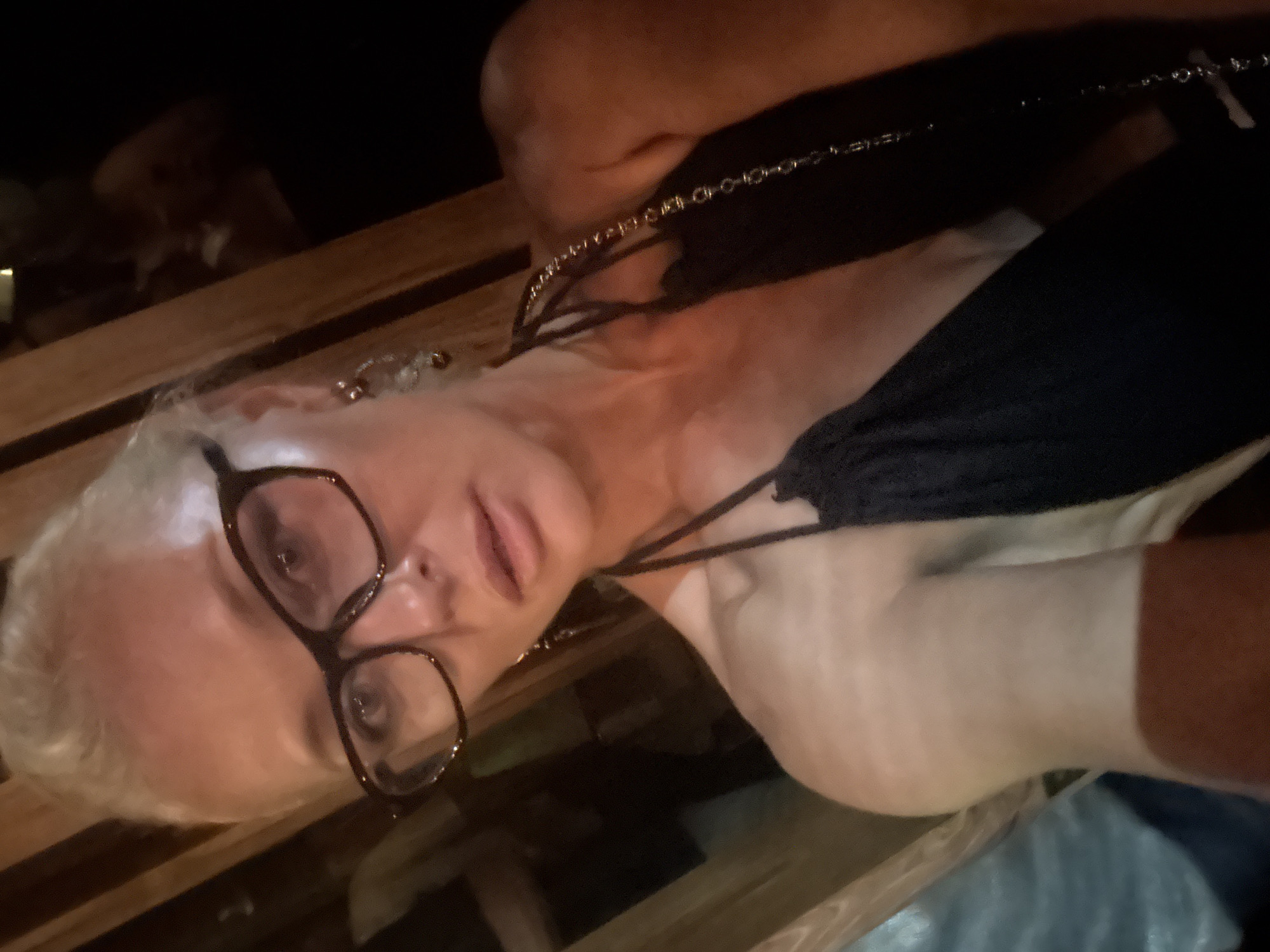 The media file is a close-up, low-angle shot of a woman with blonde hair wearing glasses. She is lying down on what appears to be a wooden surface, possibly a bench or table. Her head is tilted, and she is looking towards the camera with a relaxed expression. The lighting is dim, suggesting it might be nighttime or indoors with low light. She is wearing a dark top with thin straps and a delicate chain necklace. The background is dark and indistinct, with some blurry shapes that could be furniture or other objects. There is no visible text in the image.