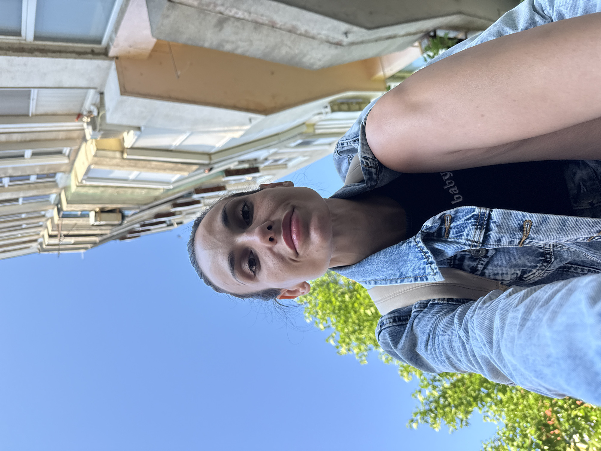 A woman takes a selfie outdoors, looking up at the camera. She is wearing a denim jacket and a black t-shirt. Behind her, there is a clear blue sky, a tree with green leaves, and a multi-story building with white windows. The building appears to be located in Pleven, Bulgaria.