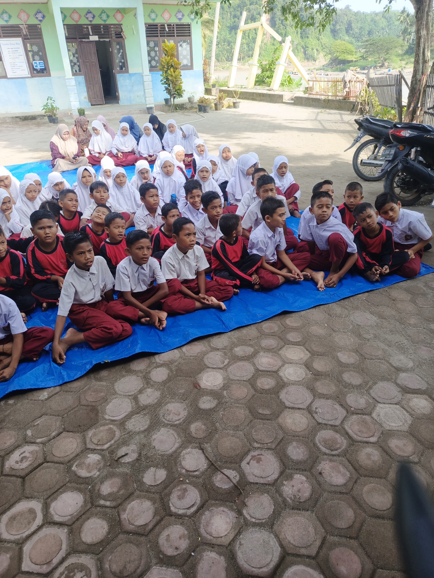 This outdoor photograph captures a group of children, likely elementary school students, and a few adults gathered on a bright, sunny day in what appears to be Simpangjaya, Indonesia. The children are predominantly wearing school uniforms; many boys are in white shirts with maroon trousers, or red and black long-sleeved shirts with maroon trousers, while many girls wear white hijabs over their uniforms, which include white tops and maroon skirts. They are all seated attentively on a large blue tarp spread over a paved area of hexagonal pavers.
In the background, a light green and blue school building with multiple windows and a brown door is visible. To the far left of the building, a whiteboard or sign displays text, with "PENJADWALAN BELAJAR SISWA" (Student Learning Schedule) clearly legible, confirming the school setting.
Beyond the paved area and the school, there is lush greenery, tall trees, and what appears to be a river or large body of water in the distance, flanked by hills. A large yellow, arch-like structure stands near the riverbank. On the right side of the image, two motorcycles are parked, and a fence runs along the edge of the paved area.
The scene suggests an outdoor class, assembly, or community gathering. The children's expressions are mostly focused and calm, indicating they are listening or observing something in the direction of the camera. The bright daylight and lack of harsh shadows suggest it is morning or early afternoon.