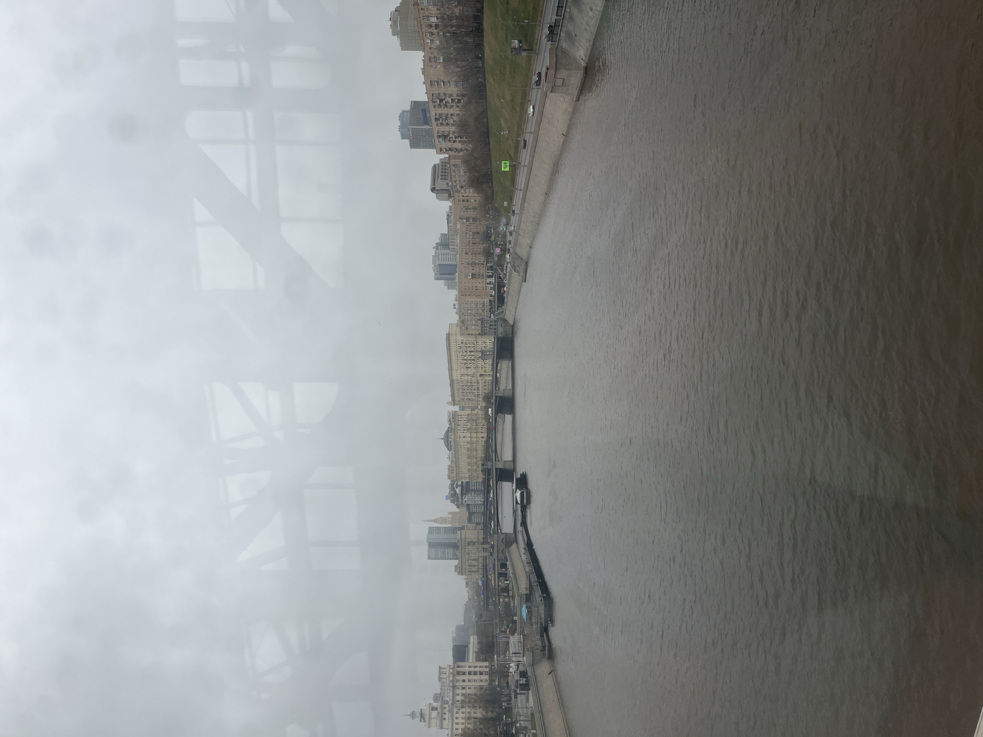 The image depicts a cityscape view, likely from a high vantage point, with a wide river dominating the right half of the frame. The river water appears murky and gray. A city skyline stretches along the left bank of the river, featuring various buildings and structures of differing heights and architectural styles. The image is taken through a window with visible framework and reflections on the left side. The sky is overcast and gray, contributing to a somber tone. The cityscape appears to be in Moscow, Russia, recognizable by the combination of historical and modern buildings. A narrow walkway or promenade runs along the riverbank. The scene appears devoid of visible people and activities.