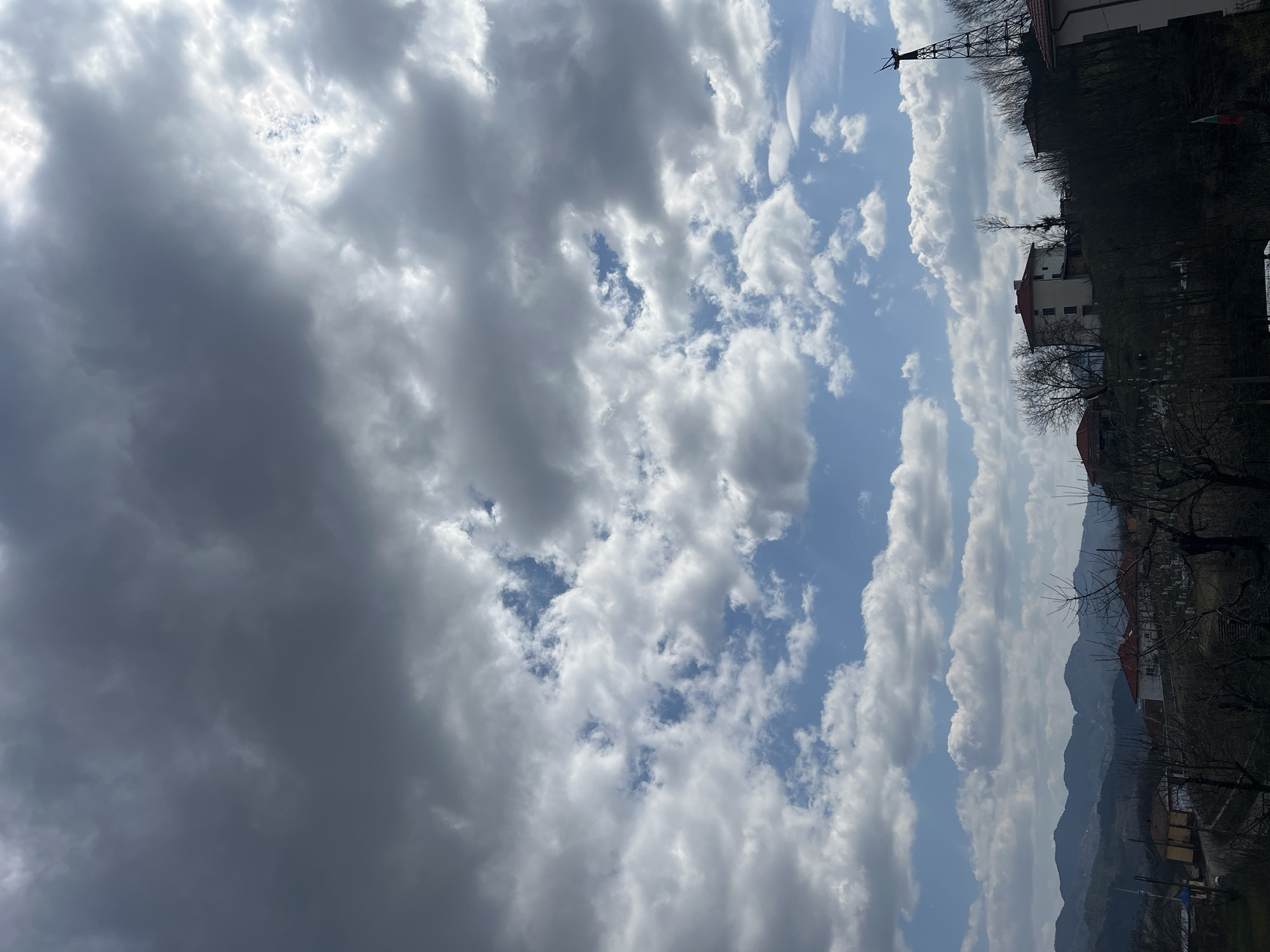 The image displays a panoramic view of Sedlartsi, Bulgaria, under a dynamic sky. The upper left portion of the sky is dominated by dense, dark grey clouds, gradually transitioning to lighter, scattered cumulus clouds across the mid-section and lower right, revealing patches of bright blue sky.
The midground features a village landscape characterized by numerous houses with predominantly red or orange tiled roofs. Several multi-story residential buildings, some painted white or light yellow, are visible, clustered on sloping terrain. A prominent feature on the right is a tall, dark, skeletal lattice tower, possibly for communication, positioned on a hill and partially obscured by bare deciduous trees, indicative of late winter or early spring. More bare trees are scattered throughout the scene, some partially obscuring views of the houses. A Bulgarian flag is subtly visible, hanging in the lower right quadrant.
In the left midground, a road or path is discernible, with outlines of parked or moving vehicles. A section of what appears to be a cemetery with numerous white markers is visible on a gentle slope in the mid-right, below some houses. The background consists of a range of hazy, undulating mountains, rendered in various shades of blue and grey, receding into the distance under the partially cloudy sky. No identifiable people or specific actions are visible within the frame.