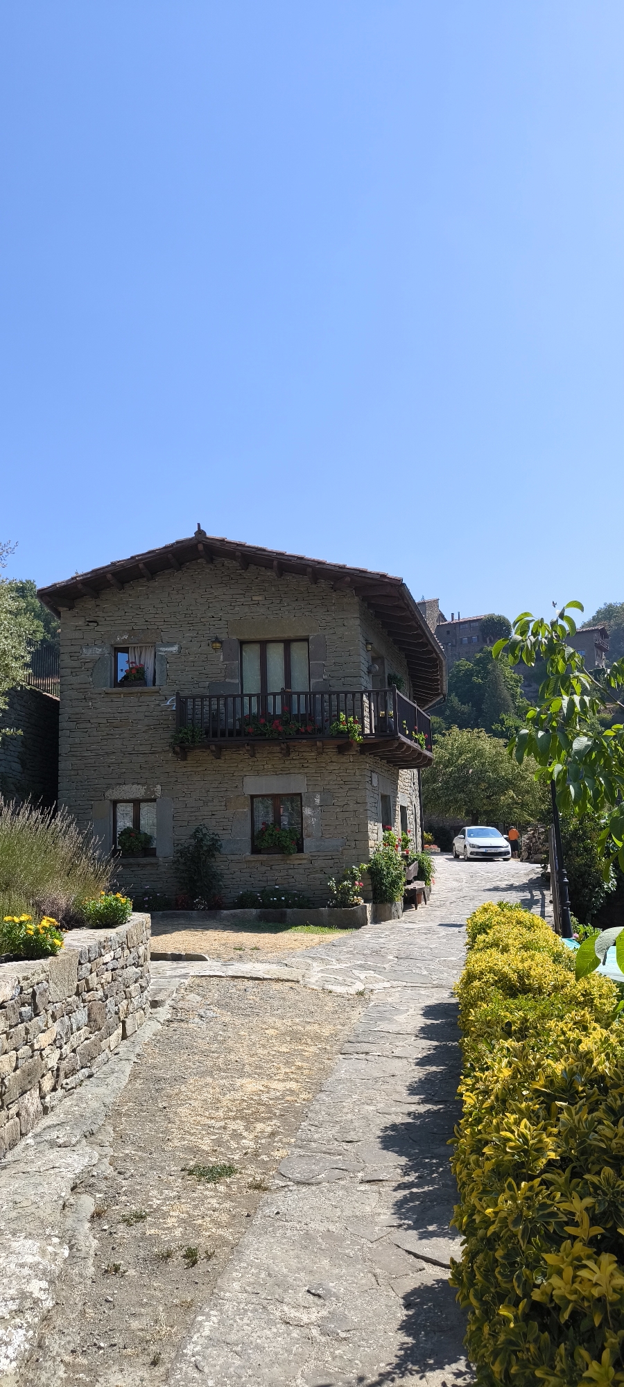 The image depicts a sunlit, outdoor scene in the village of Rupit i Pruit, Spain. In the foreground, a stone pathway leads towards a two-story stone building. The building has a rustic appearance with a tiled roof and wooden balcony adorned with red flowers. Several windows with flower boxes are visible. To the left of the pathway, a low stone wall is covered in yellow and green foliage, with lavender bushes and more yellow flowers. To the right of the building, the pathway curves and continues uphill, flanked by a dense hedge of yellow-green bushes. Further up the path, a white car is parked, and a person in an orange shirt is visible near it. In the background, more stone buildings and lush green trees are scattered across a hillside under a clear blue sky. The overall atmosphere is tranquil and picturesque, characteristic of a Mediterranean village.