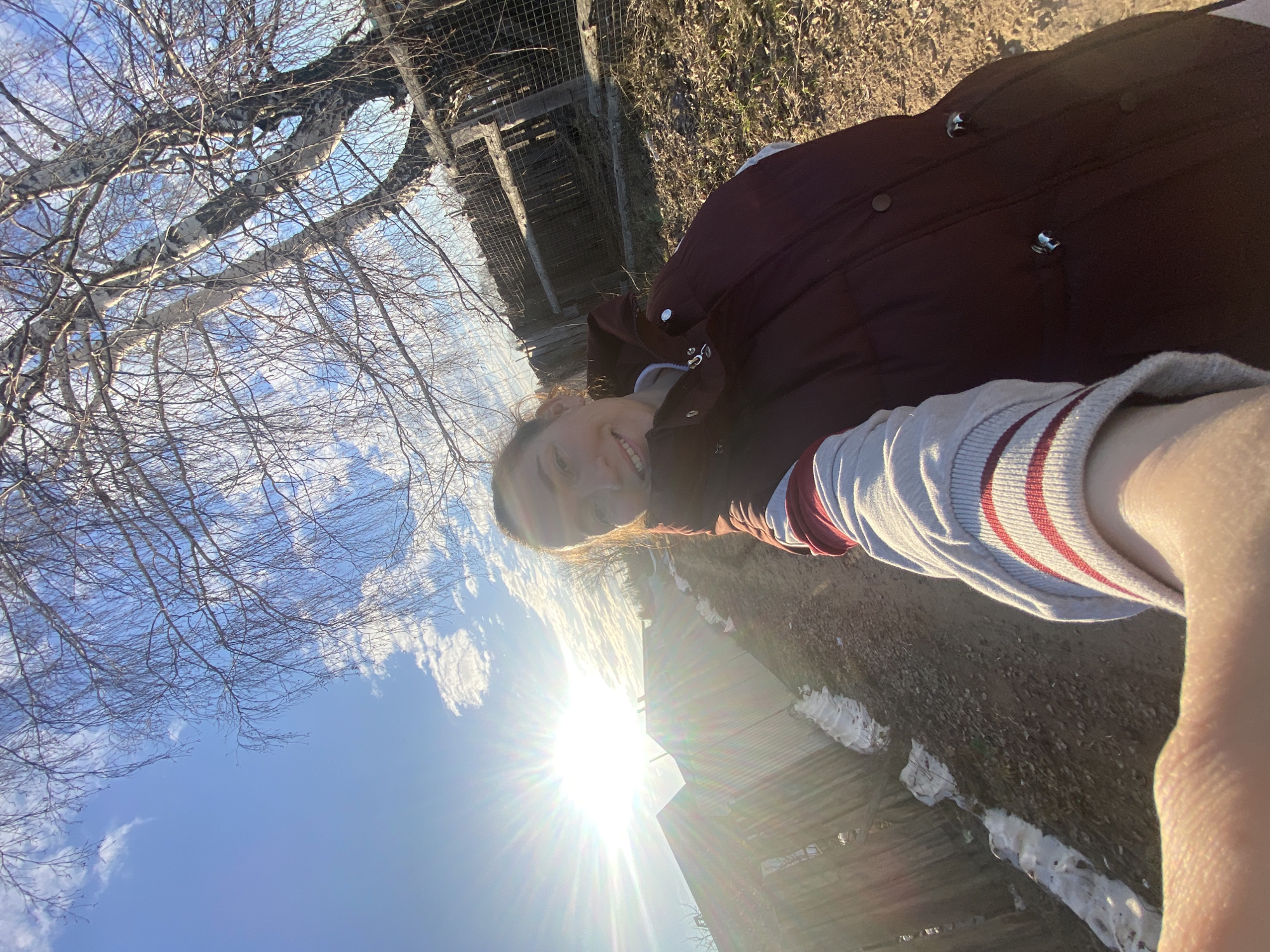 A photograph, taken in landscape orientation, depicts a person outdoors. The background features bare tree branches against a blue sky with wisps of white clouds. A wooden fence-like structure is visible behind the person. Patches of snow are present on the ground. The sun is shining brightly, casting a lens flare and creating a high-key lighting effect. The photograph was likely taken in Ольховка, Russia.
