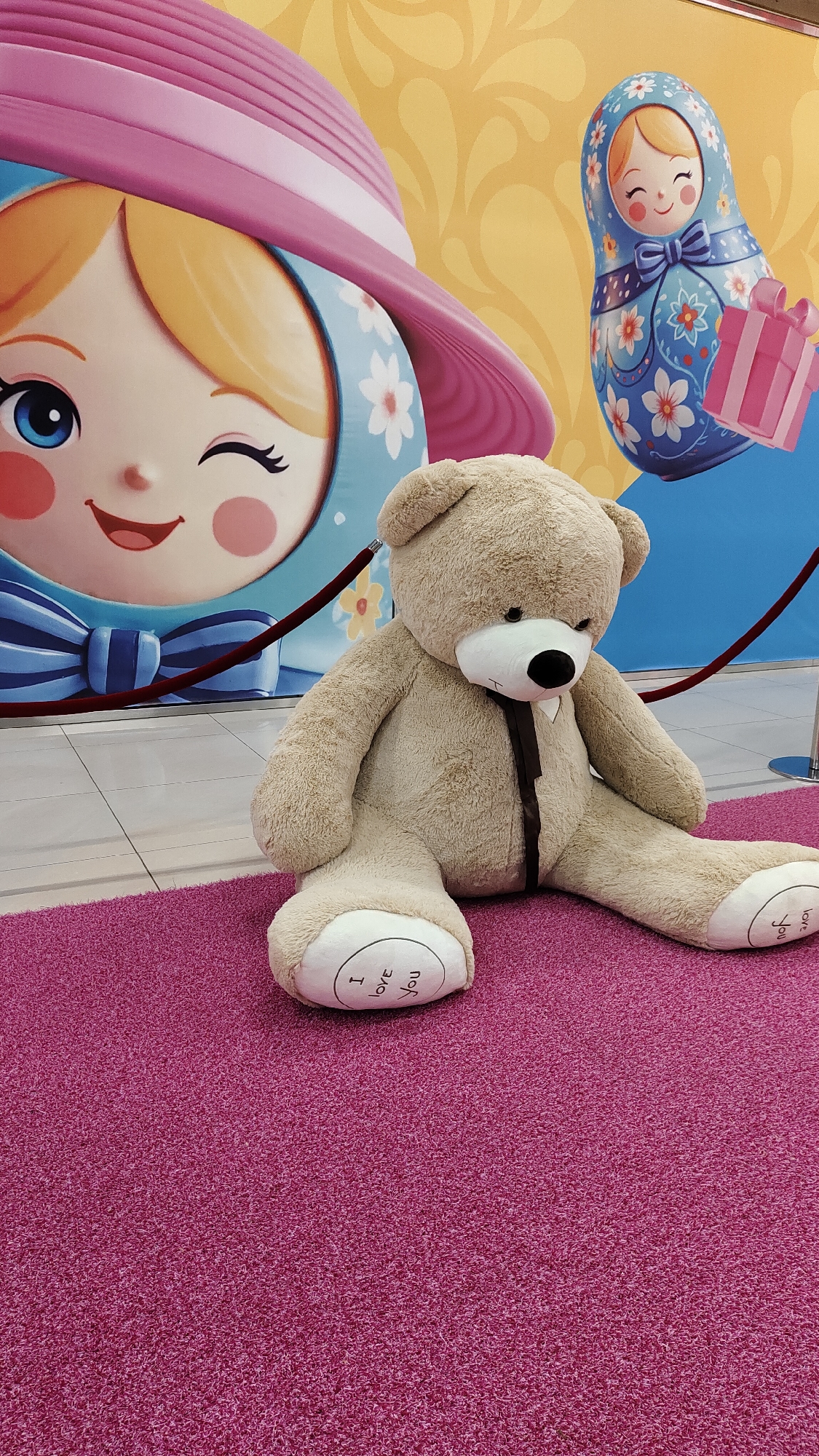 An oversized, light brown teddy bear sits on a pink carpet in front of a large mural. The mural depicts two Matryoshka dolls, one on the left with a large pink hat and a blue and white floral pattern on its dress, and another on the right that is entirely blue and white with a floral pattern and a pink gift box. The teddy bear is in a seated position with its legs extended. On its left foot, the words "I love you" are printed in black cursive text. On its right foot, the words "I love you" are also printed. Red velvet ropes are visible in the background, indicating a barrier. The floor beneath the carpet appears to be tiled. The environment suggests an indoor public space, possibly a shopping mall or an event venue.