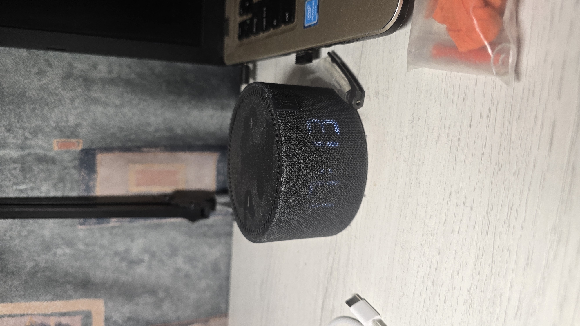 The image shows a close-up of a black smart speaker placed on a white wooden surface. The smart speaker is cylindrical and has a mesh fabric covering. A blue LED display on the speaker shows the time "13:11". The speaker appears to be connected to a laptop, which is visible in the upper portion of the image. The laptop is silver and has a black keyboard. A portion of a clear plastic bag containing orange and red objects is visible to the right of the speaker. In the background, a patterned surface, possibly a rug or wallpaper, can be seen. The overall scene suggests a workspace or a desk setup. There is no visible indication of time of day, weather, or emotions. The visible text on the laptop includes the Intel logo.