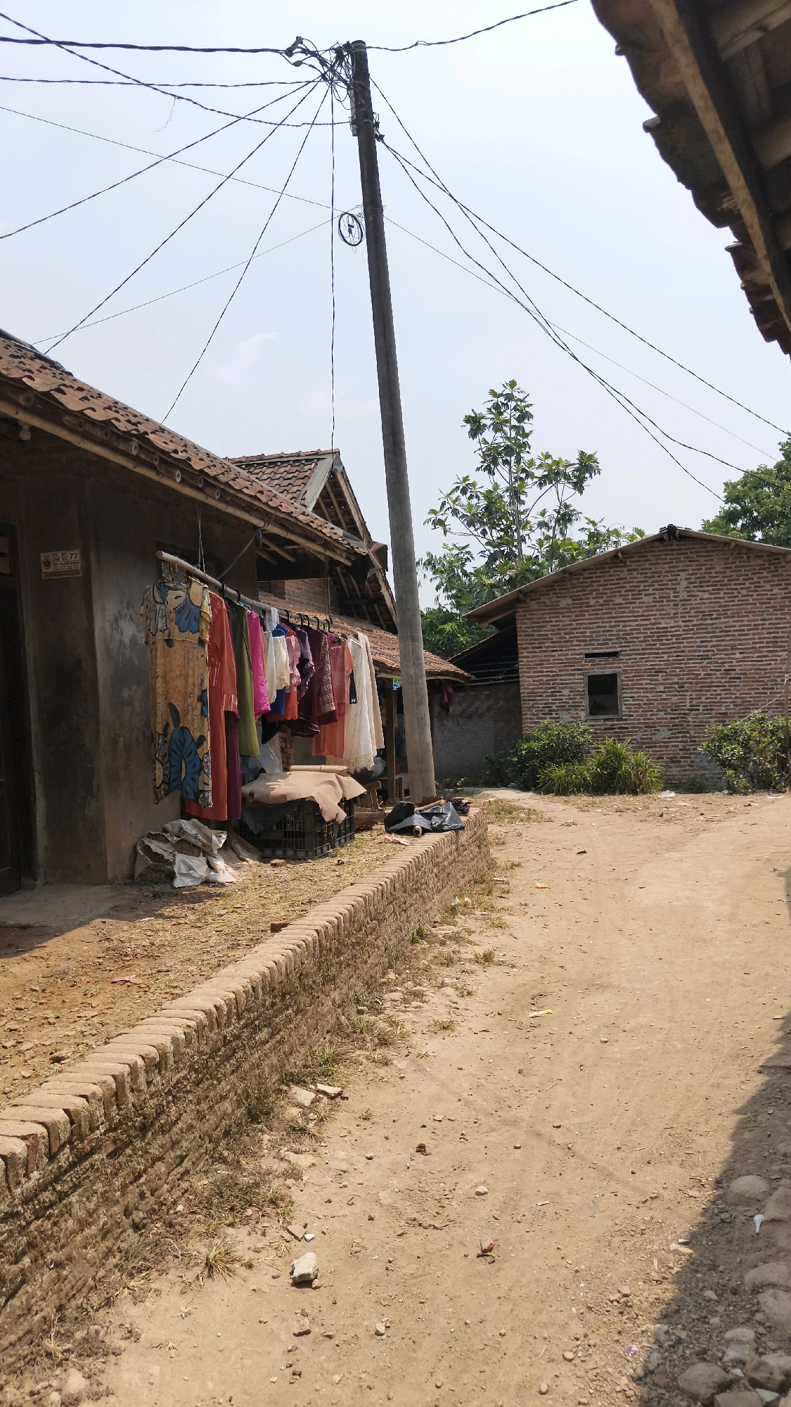 In a dusty, sunlit outdoor scene, likely in the city of Tenjo, Indonesia, a rustic pathway is bordered by a low brick wall. To the left, a simple concrete house features a clothesline hung with colorful garments. Adjacent to this house, a second brick structure stands with a small, dark window. A tall, weathered utility pole with numerous tangled wires stretches into the bright, clear sky. Trees and foliage dot the background. The overall impression is one of a rural or semi-rural residential area under a bright, sunny sky.