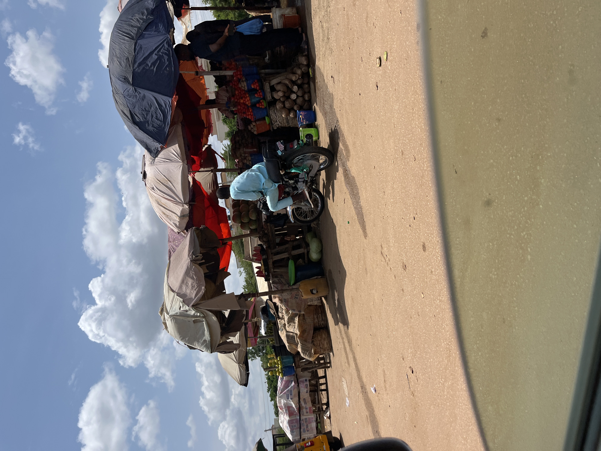 The image shows a street scene in Galadimawa Village, Nigeria. A row of market stalls lines the left side of the road, shaded by numerous umbrellas. The stalls display various goods, including fruits, vegetables, and firewood. A man in a light blue shirt sits on a motorcycle, appearing to browse the stalls. The road is paved and extends into the distance. The sky is visible on the left, with scattered clouds. The image appears to be taken from inside a vehicle, as part of the dashboard is visible in the foreground on the right.