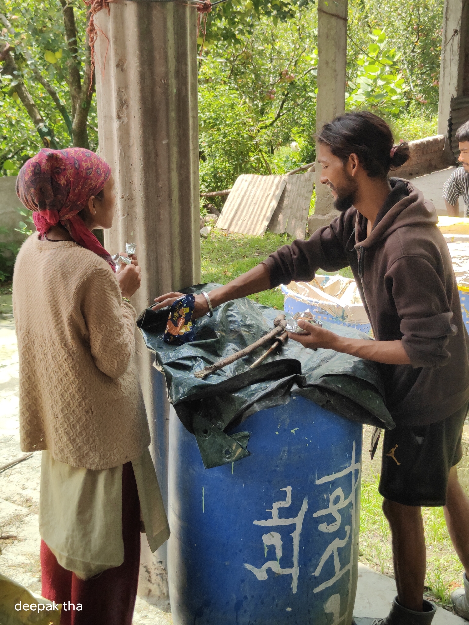 Two individuals are present in an outdoor setting, possibly in a rural or semi-urban area, with lush green foliage and trees in the background. The scene appears to be in Baran, India, suggested by the watermark "deepak tha."
On the left, a person wearing a red and pink patterned headscarf, a beige patterned sweater, and a lighter-colored skirt over red leggings stands facing a large blue barrel. They are holding a small, clear plastic object in their right hand.
On the right, a person with dark hair tied back in a bun, wearing a brown hooded sweatshirt and dark shorts with a "Jordan" logo, is leaning over the blue barrel. They are interacting with the barrel and seem to be handing or receiving something from the person on the left. Their left hand is extended towards the barrel, and their right hand is near a small, metallic object.
The blue barrel is covered with a dark green tarp. Several pieces of debris, including wooden sticks and what appears to be a piece of folded fabric with an image on it, are visible on the tarp. The barrel itself has white markings that resemble script or symbols. A corrugated metal sheet is leaning against a concrete pillar in the background, and another person is partially visible on the far right, wearing a striped shirt. The ground is covered with dirt and some grass. The lighting suggests it is daytime.