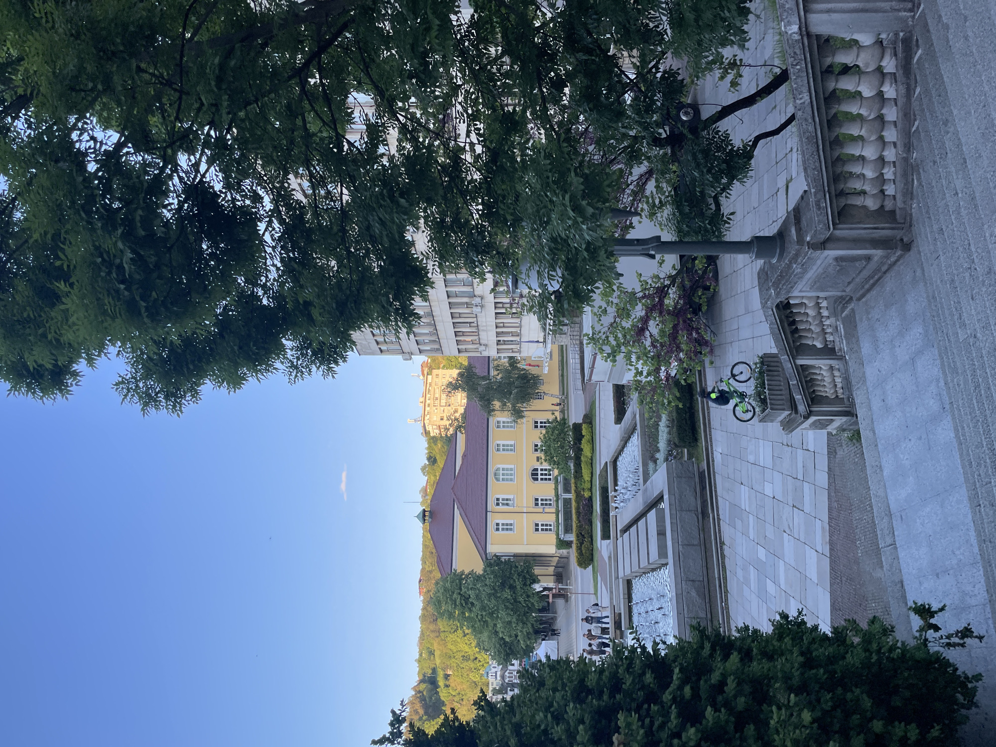 The photograph captures a scene in Gabrovo, Bulgaria, featuring a view overlooking a city square or park area. The dominant structure is a yellow building with a red roof and white-framed windows. The building is surrounded by greenery, including trees, and nestled against a hillside.
In the foreground, there are stone-paved areas with what appear to be shallow reflecting pools or fountains with water features. A few people are visible in the distance. A green bicycle is resting against the railing. The sky is clear blue, with tree foliage framing the upper portion of the image. The perspective is slightly elevated, suggesting the photo was taken from a higher position overlooking the square.