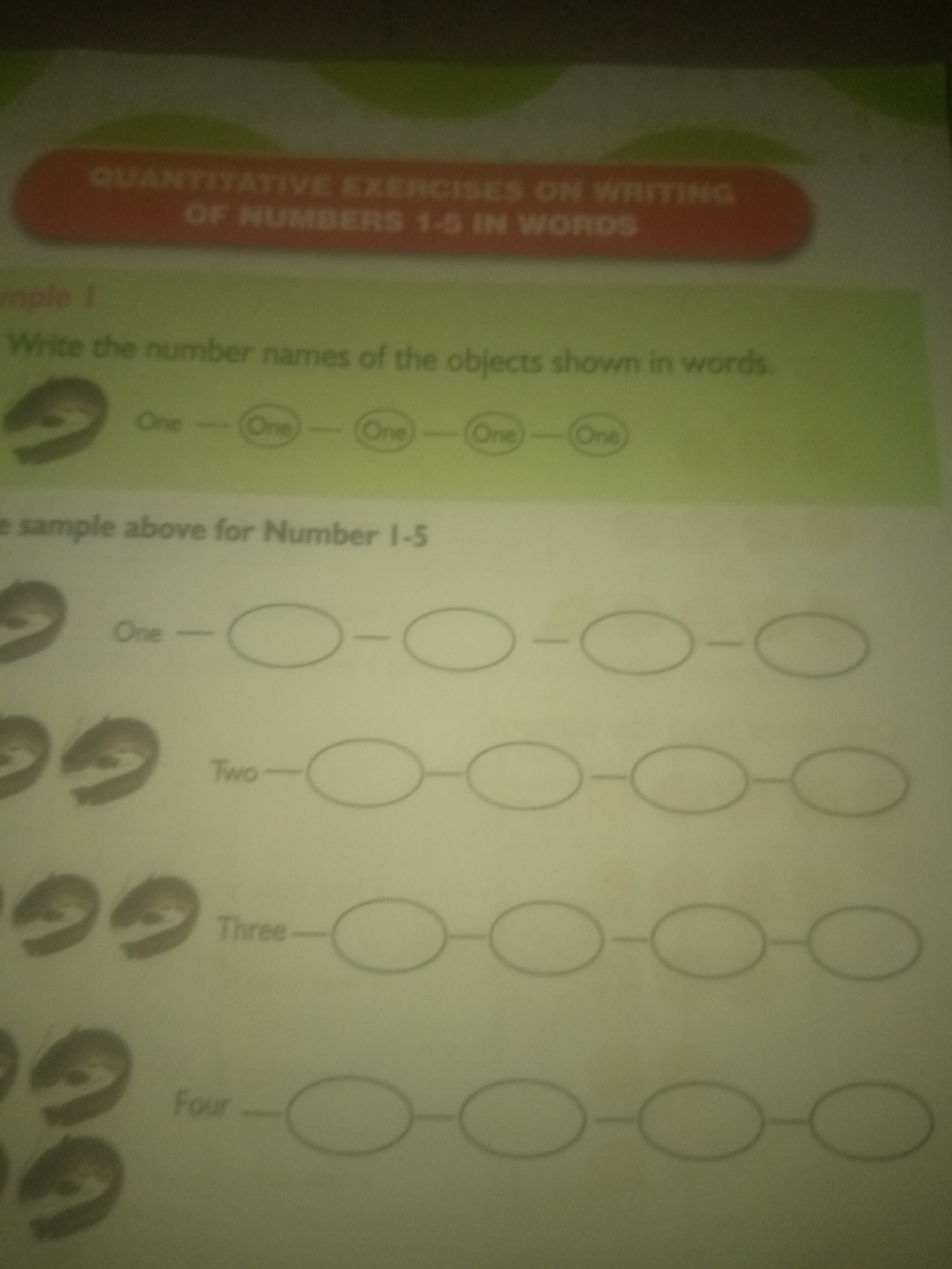 The image shows a page from a workbook or educational material, likely for young children learning to write numbers. At the top, a banner reads "QUANTITATIVE EXERCISES ON WRITING OF NUMBERS 1-5 IN WORDS". Below this is an example labeled "Example 1" which shows a drawing of an object followed by the word "One" and then four oval shapes, each containing the word "One" within it, connected by hyphens.
The main section of the page shows a grid-like layout with numbers written on the left, followed by a series of oval shapes connected by hyphens.
The first row has the number "One" written next to four empty oval shapes.
The second row has the number "Two" written next to three empty oval shapes.
The third row has the number "Three" written next to three empty oval shapes.
The fourth row has the number "Four" written next to three empty oval shapes.
There are also images of small, cartoon-like faces or figures on the left side of each row, corresponding to the number of ovals in that row, although they are partially cut off at the top of the image. These figures appear to be identical in each row.
The image quality is somewhat low, and the lighting is uneven, suggesting it was taken with a camera in a room with artificial light. There is no discernible connection to Jauro Yinu, Nigeria. The content is purely educational.