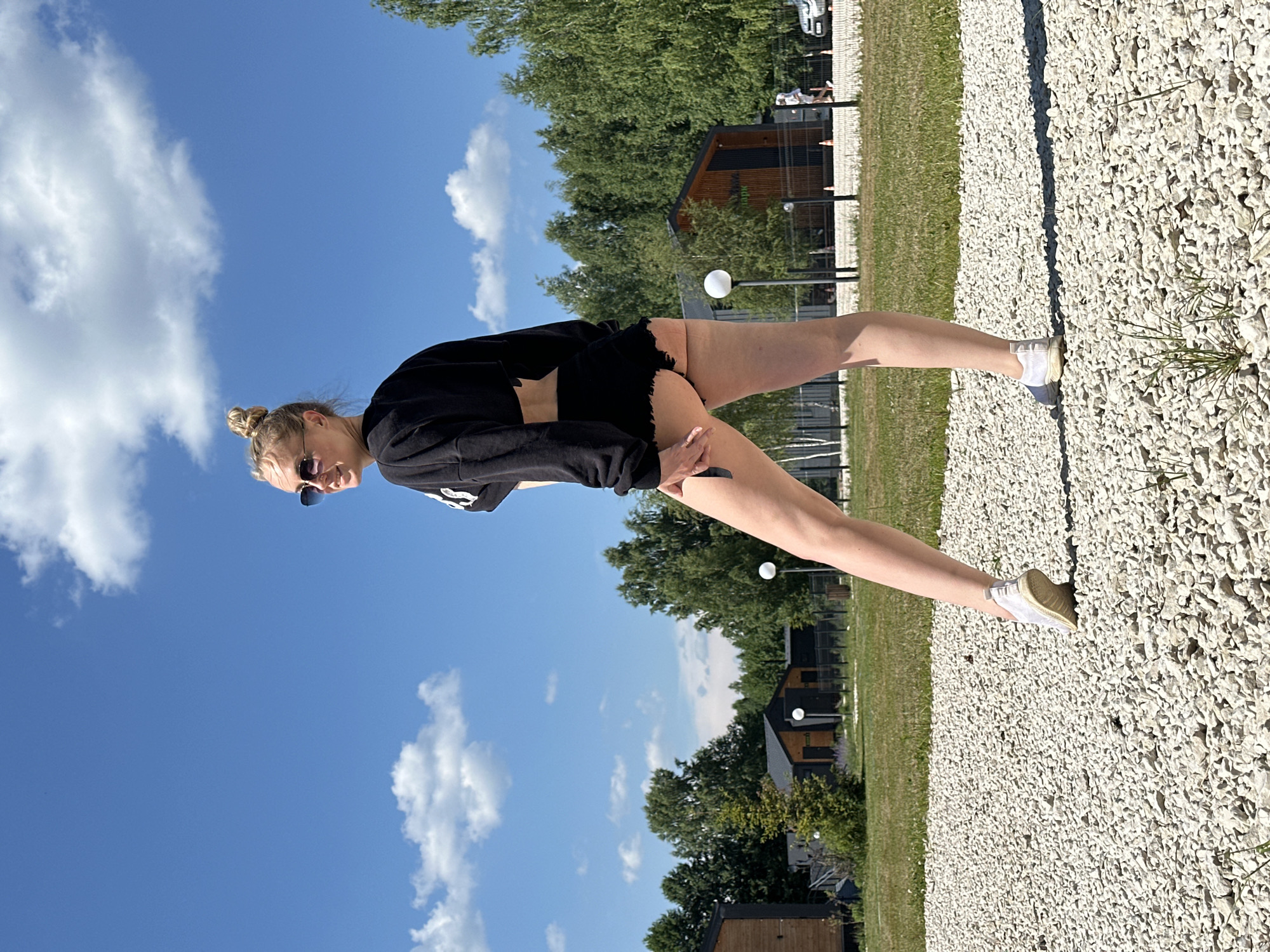 A young woman with blonde hair in a bun, wearing sunglasses, a black crop top, and black shorts, stands in a gravel area. She is looking over her left shoulder towards the camera. Behind her, there is a grassy area with several modern, wooden houses and a line of trees. The sky is blue with scattered white clouds. The image appears to be taken outdoors in a recreational or residential area.