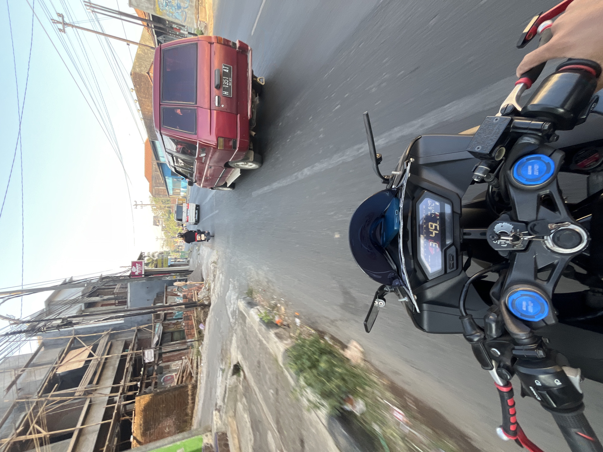 The image shows a first-person view from a motorcycle as it travels down a street in Pecangaan Wetan, Indonesia. The rider's hands are visible on the handlebars, and the motorcycle's digital dashboard displays "3:49". To the right, a red van with license plate "H 1351 AT" is in motion. Further down the road, another white van is visible. On the left side of the street, a building is under construction with scaffolding, and the sidewalk is lined with debris. The sky is clear and blue, indicating a daytime scene.