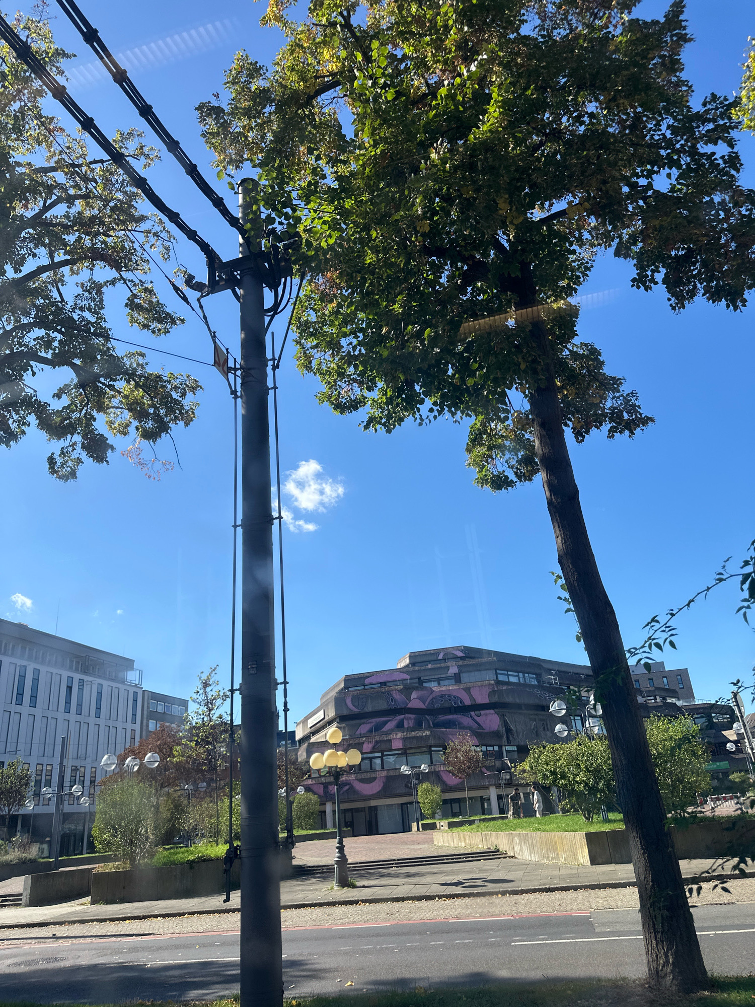 The image shows a street scene with a distinctive building featuring purple art on its facade. The building is a Brutalist-style structure with multi-levelled, angular sections. In front of the building, there is a park-like area with trees, grass, and concrete planters. A lamppost with multiple yellow lights stands in this area.
To the left of the building, a modern, multi-story building with a white facade and numerous windows is visible. A utility pole with cables runs vertically through the center of the image, obscuring parts of the view. Trees with green and yellowing leaves are prominent, some of them partially obscuring the sky. The sky is blue with a few small white clouds.
A paved road is in the foreground, with a sidewalk and a low concrete wall separating it from the park area. There are two individuals standing on the grass in the middle ground. The scene suggests an urban environment, likely in a city due to the architecture and infrastructure.
Based on the architectural style and the presence of the specific building, this location is identified as the **Krefeld University of Applied Sciences (Hochschule Niederrhein)** in Krefeld, Germany. The building with the purple art is the **"Die Eule"** (The Owl) building, a well-known structure on the campus.