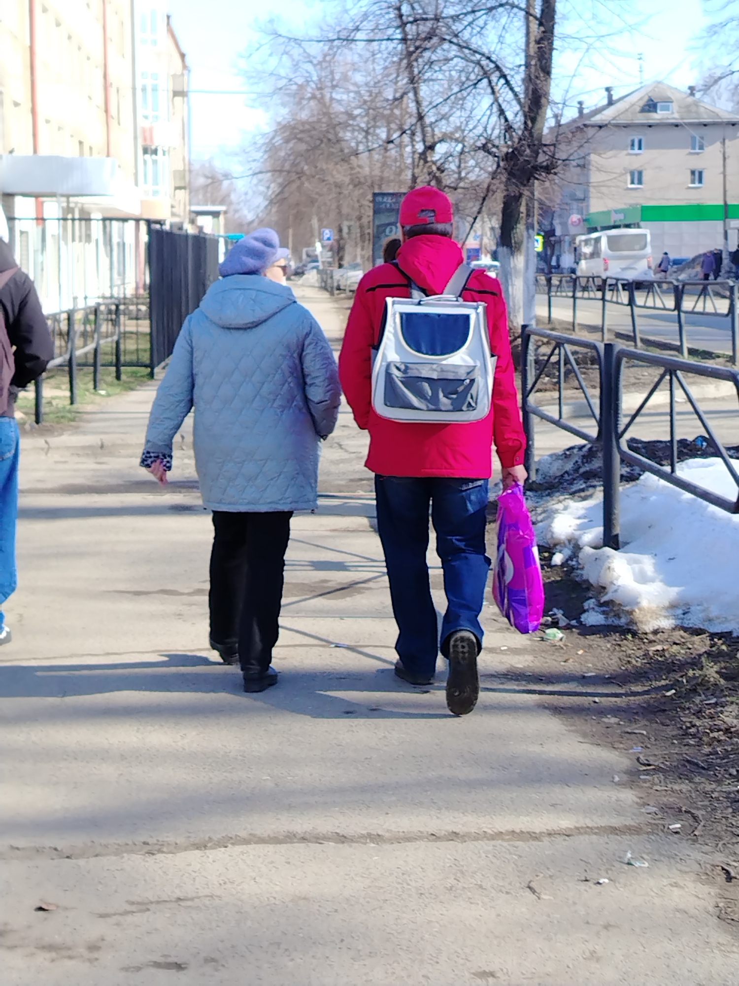 The image shows two people walking on a sidewalk in the city of Chaikovsky, Russia. A person wearing a red jacket, blue jeans, and a red cap is walking in front, carrying a purple plastic bag and a backpack. An older person wearing a blue quilted jacket and black pants is walking behind the first person. The sidewalk is next to a street with a black metal fence, and there is some snow on the ground. Several buildings are visible in the background.