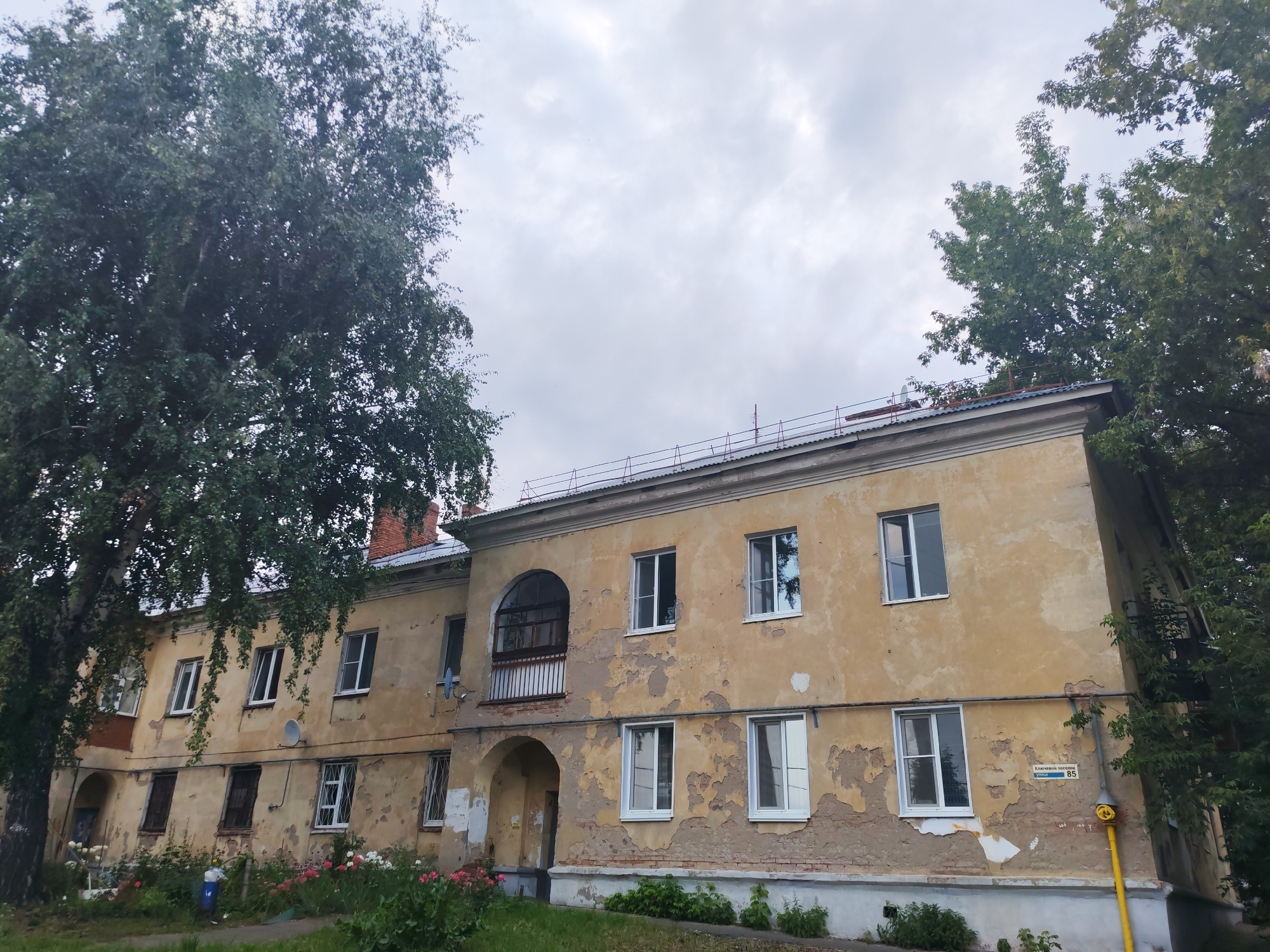 The image shows a dilapidated, yellow-painted apartment building in Izhevsk, Russia, under a cloudy sky. The building has peeling paint, revealing patches of brickwork beneath. There are numerous windows, some with white frames and others with dark grates. A balcony with a dark railing is visible on the second floor, above an arched entryway. A large, leafy tree obscures part of the building on the left, and another tree is visible on the right. In the foreground, there is a patch of green grass with some flowers and a blue trash can. A yellow gas pipe runs vertically along the right side of the building. A street sign indicating "Ключенацкая переулок 85" (Klyuchenatskaya Lane 85) is affixed to the wall. The overall scene suggests an older residential area in need of repair.