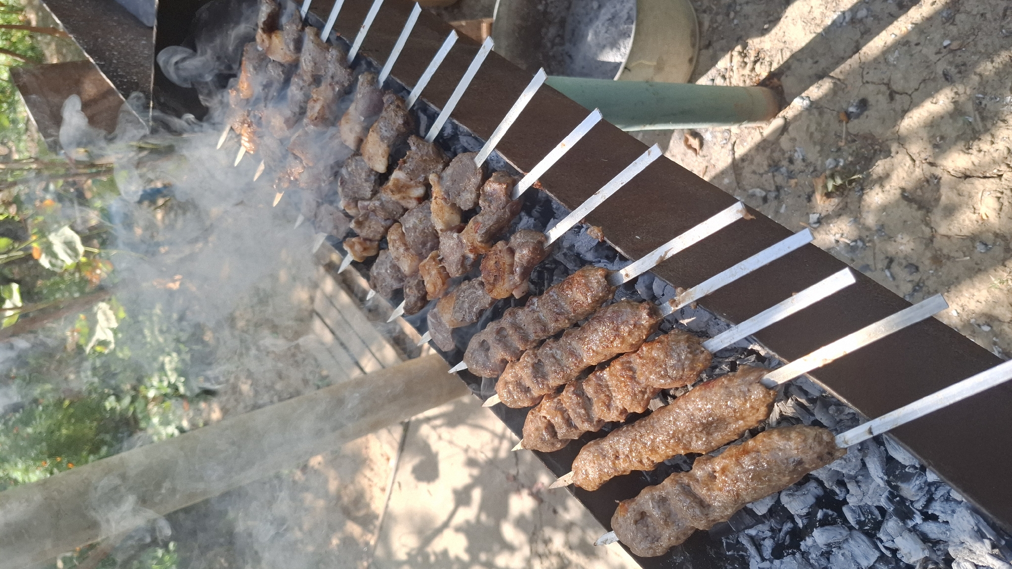 In the city of Шұбарсу, Kazakhstan, an outdoor barbecue is underway. Several skewers of meat, some appearing to be ground meat formed into a long shape and others cubed, are being grilled over hot coals. Smoke rises from the barbecue, indicating the cooking process. The barbecue is situated on a dirt surface, with scattered debris and shadows from nearby foliage. In the background, greenery and trees are visible, suggesting a natural or semi-natural outdoor setting. The scene is bathed in sunlight.