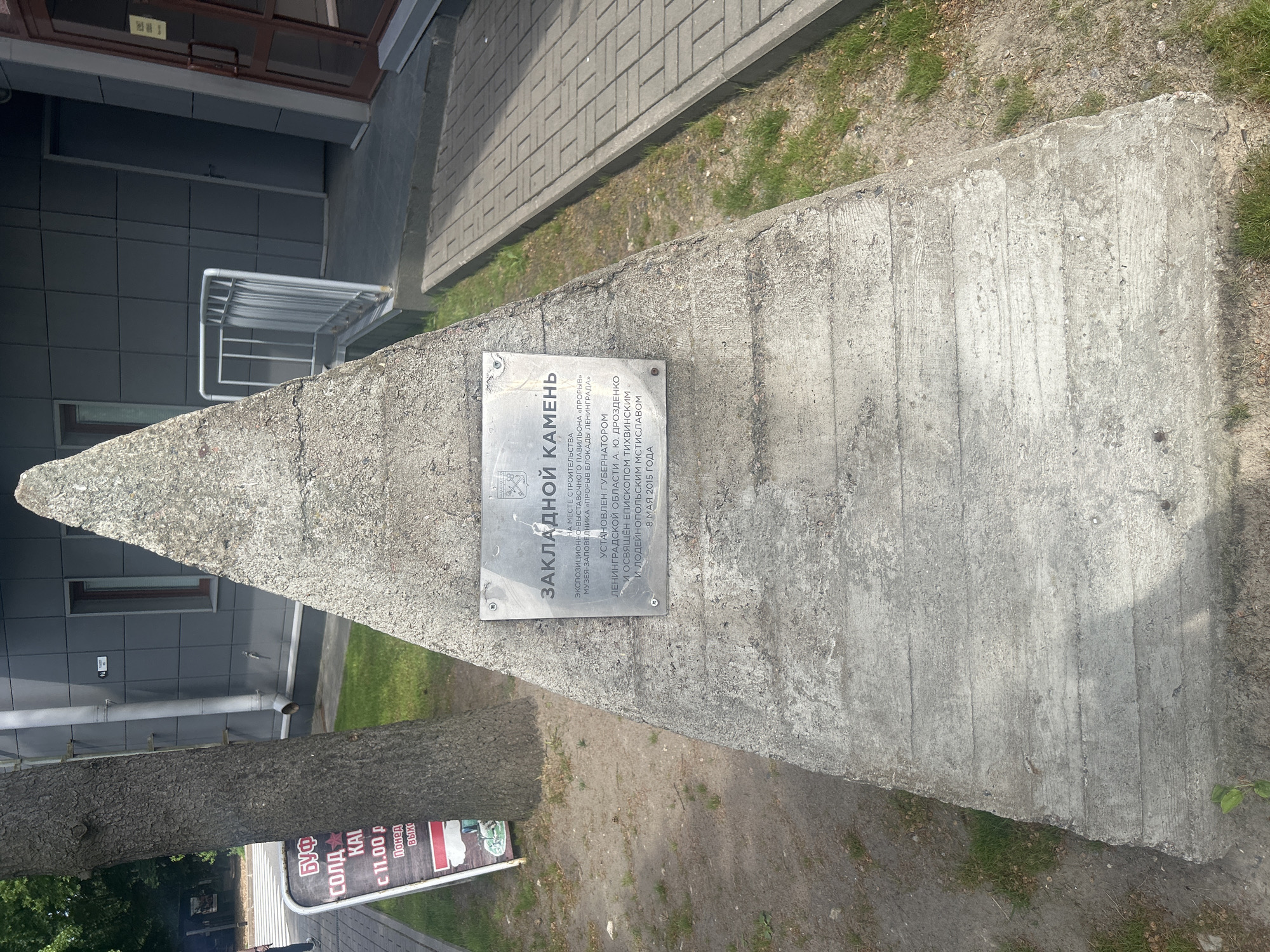 A concrete pyramid-shaped monument stands on a paved area in front of a modern building with grey tiled walls and a glass door. A metal plaque is affixed to the monument. A tree trunk is to the left of the monument. A sidewalk with brick-like paving extends from the foreground to the right of the image. The monument has a plaque that reads "ЗАКЛАДНОЙ КАМЕНЬ" (Founding Stone) and lists the location as "НА МЕСТЕ СТРОИТЕЛЬСТВА ЭКСПОЗИЦИОННО-ВЫСТАВОЧНОГО МУЗЕЯ-ЗАПОВЕДНИКА «ПРОРЫВ» БЛОКАДЫ ЛЕНИНГРАДА" (At the site of construction of the exhibition-museum complex "Breakthrough" of the Siege of Leningrad). It also states it was installed by Governor A. Yu. Drozdenko of Leningrad Oblast and consecrated by Bishop Tikhvin and Ludinopolislav Mstislav on May 8, 2015. This monument is located in Shlisselburg, Russia, specifically at the site of the "Breakthrough" Museum.