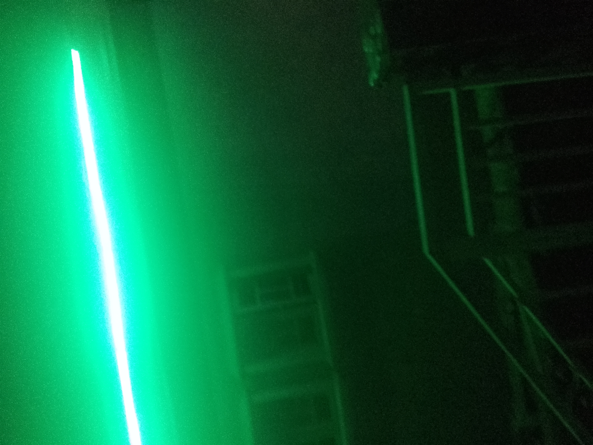 The image depicts a dimly lit outdoor scene, likely at night, dominated by a bright, vertical green light source on the left side. This light casts an intense glow, illuminating the textured surface it's projected onto, which appears to be a wall. The right side of the image is darker, revealing structural elements that resemble a staircase or scaffolding against a dark background. Faint, rectangular shapes, possibly windows or architectural features, are visible on a building in the mid-ground, partially obscured by the ambient green light and shadows. The overall atmosphere is abstract and somewhat mysterious due to the limited visibility and the strong monochromatic lighting. The provided information indicates this scene is in Jalingo, Nigeria, but the visual content itself does not offer specific geographic markers to confirm this location beyond the metadata.
