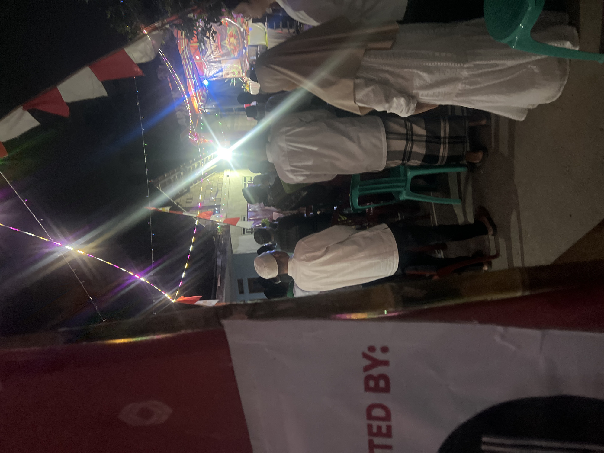 The image captures a nighttime outdoor gathering in Klapanunggal, Indonesia, likely a community event. In the foreground, a partial view of a red and white banner displays the text "TED BY:". Several individuals are visible, most dressed in traditional Indonesian attire, including white shirts and peci caps for men, and a white hijab and dress for a woman. They appear to be standing and observing something, possibly a stage or performance area in the background. The background is illuminated by a series of colorful string lights and a bright spotlight, suggesting a stage setup with a banner that reads "Festival" and "Kampung". Red and white Indonesian flags are also present, draped along the string lights. The environment appears to be a street or open area, with a rough concrete ground. The overall atmosphere is festive, typical of local celebrations in the region.