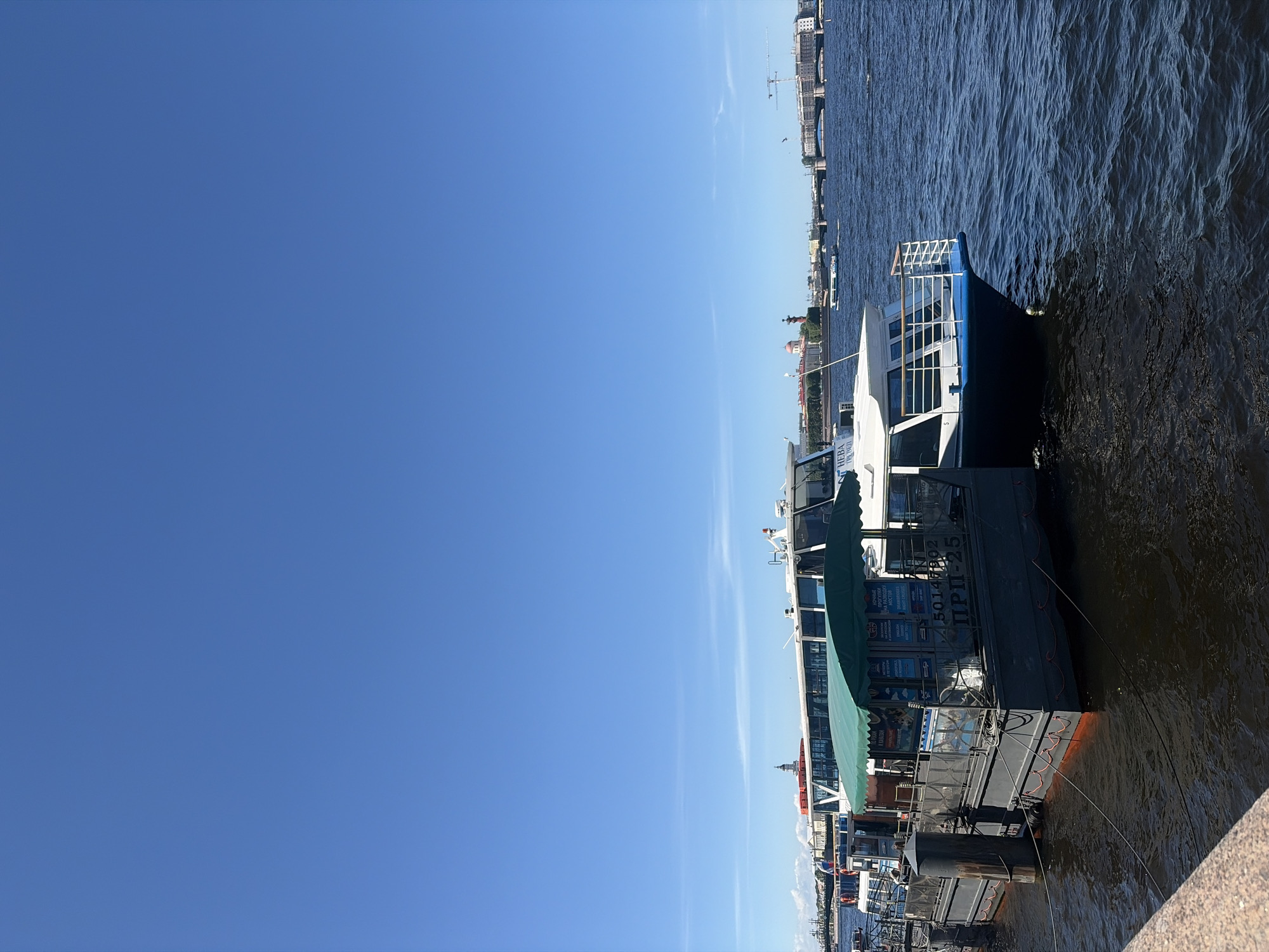 This detailed image captures a daytime scene along the Neva River in Saint Petersburg, Russia, featuring moored boats and the city's architectural panorama under a clear sky.
In the foreground, a light-colored, rough-textured stone embankment is visible along the bottom edge. Extending into the water from the left, a dark grey, rectangular floating platform or dock is securely moored to two dark, cylindrical bollards by thick lines. This platform features a green scalloped awning overhead, providing shade. Its side panels display various informational signs in both Russian and English, advertising boat tours, including "НОЧНЫЕ ЭКСКУРСИИ НА РАЗВОД МОСТОВ" (Night excursions to bridge openings) and "ПО НЕВЕ И КАНАЛАМ" (Along the Neva and canals), as well as "ENGLISH SPEAKING BOAT TOURS". The numbers "5014902" and the identifier "ПРП-25" are stenciled on the side of the floating structure.
To the right of the floating platform, a white and dark blue passenger boat, likely a tour vessel, is moored. Its bow is prominent, featuring white railings and large, rectangular windows. A partial name, "НЕВА ТРЕВЕЛ," is visible on its hull. The water around the boats is dark blue-grey, exhibiting ripples.
In the midground, the Neva River stretches across the frame, with another smaller, light-colored boat actively traversing the water.
The background presents a distinctive Saint Petersburg skyline. Prominent landmarks visible on the opposite bank include a red-colored building with a white dome, characteristic of structures on Vasilievsky Island, potentially near the Rostral Columns, one of which is clearly discernible as a tall, slender red column with white sculptural details. Multiple bridges span the river in the distance. Further to the right, beyond the bridges, modern buildings and at least two construction cranes indicate ongoing development.
The sky occupies the upper half of the image, displaying a vivid, uniform blue with only a few faint, wispy white cloud trails or contrails visible at high altitude. The scene is brightly illuminated by direct sunlight, casting clear shadows and highlighting the details of the boats and distant architecture. No individuals are distinctly visible in the immediate foreground, though passengers may be present within the moored tour boats.