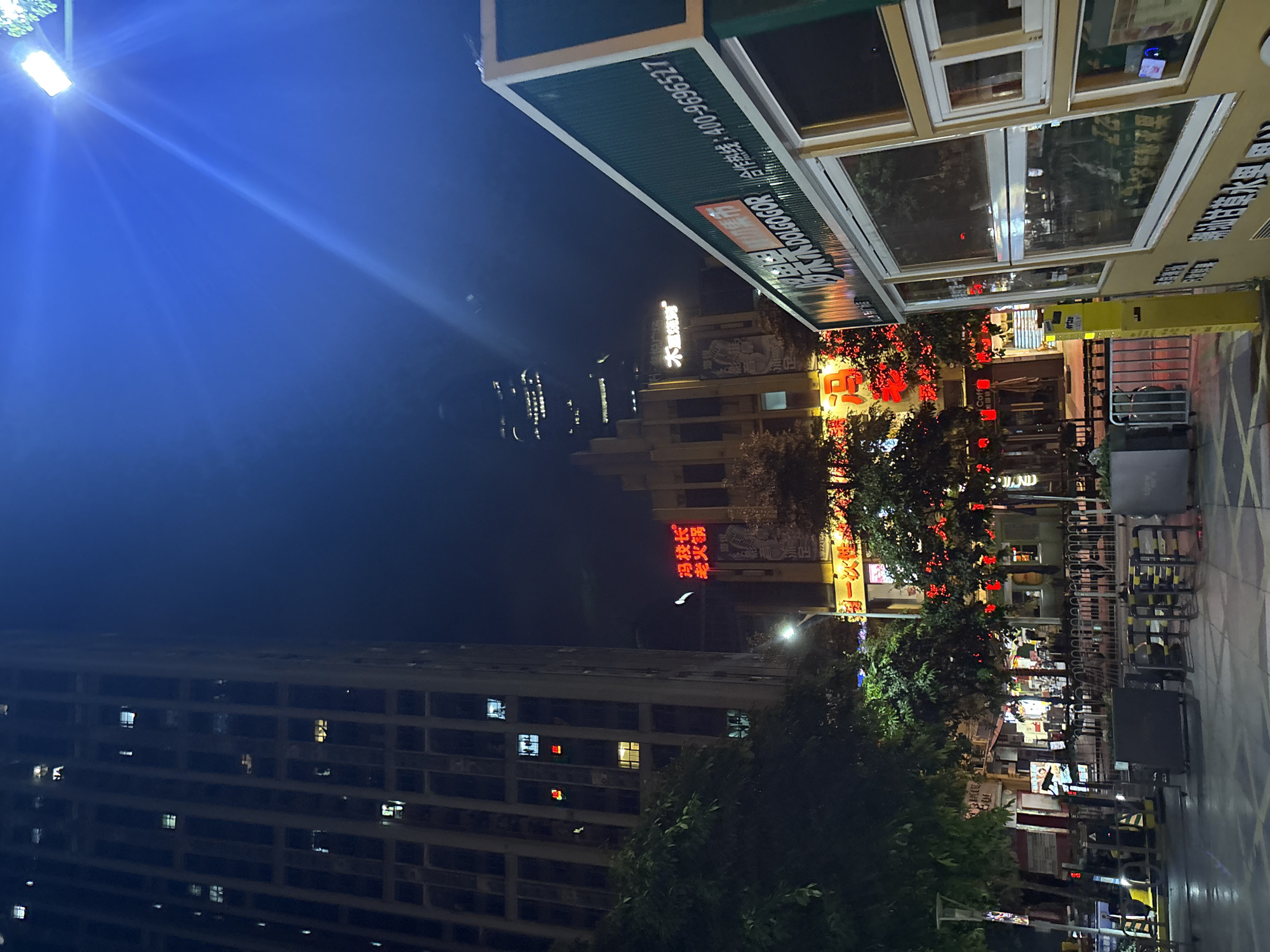This is a nighttime, eye-level, outdoor shot of a street scene in Haizhu District, China. The image is vertically oriented. A tall apartment building dominates the left side of the frame, its windows illuminated with warm light. To the right of the apartment building is a street lined with shops and trees. Red lanterns and neon signs cast a vibrant glow on the scene. Text in the image includes "冯校长," "老火锅," "木風烧烤," "Coffe," and "ertze." A prominent green sign displays "木木DOLGOGOR 合作热线:400-9696527," which appears to be a hotline number. There are several metal barriers and a cart on the right side of the street, suggesting a commercial or pedestrian area. The sky is dark blue, indicating it is nighttime. The overall atmosphere is lively and bustling, typical of a commercial street after dark. No people are clearly visible.