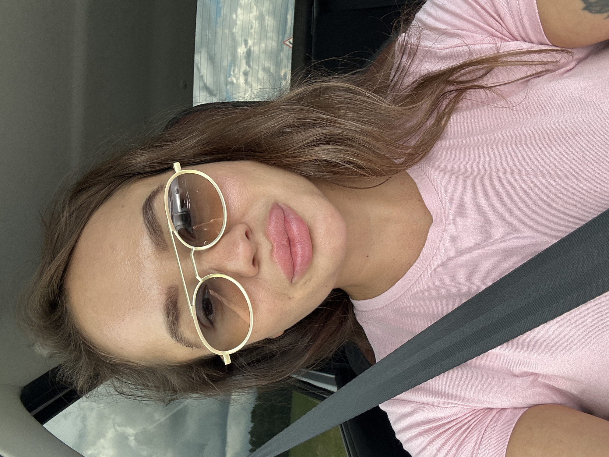 A young woman with light brown hair is sitting in the driver's seat of a car, facing the camera. She is wearing a pink t-shirt and gold-rimmed sunglasses. A gray seatbelt is visible diagonally across her chest. The background shows a slightly overcast sky and greenery through the car windows, suggesting an outdoor location, possibly rural or on the outskirts of Uvel'skiy, Russia.