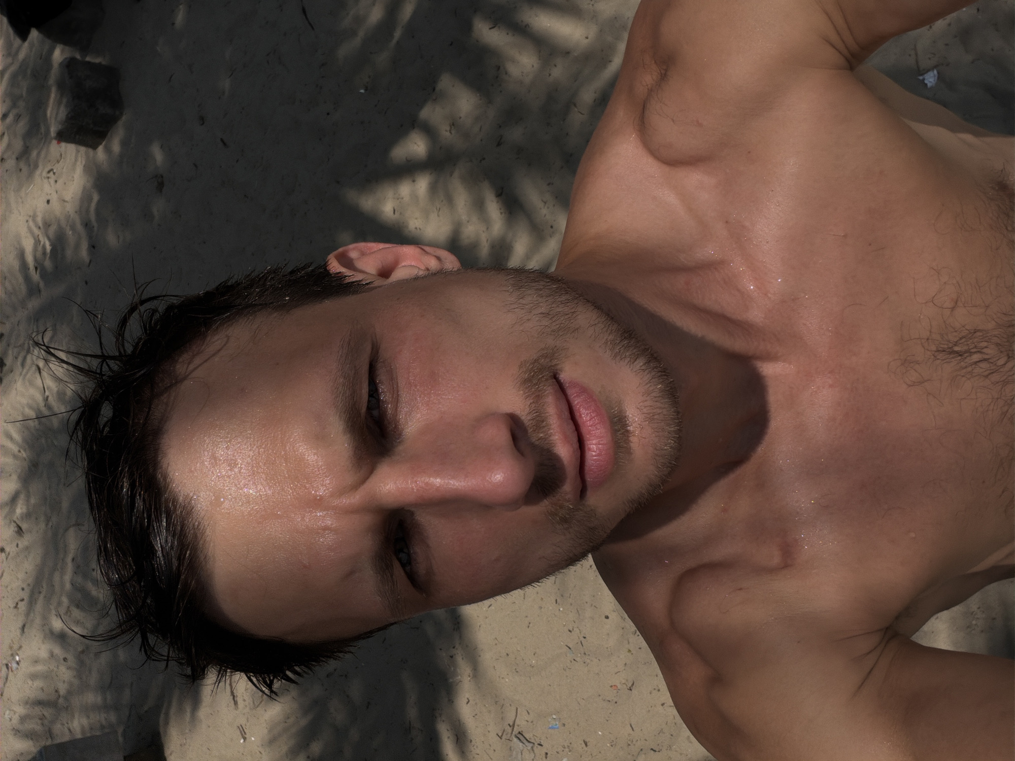 A sun-drenched, close-up shot depicts a man lying on sandy ground. His face is turned towards the camera, revealing wet hair, a light beard, and slightly parted lips. The man's chest and part of his upper body are visible, with droplets of moisture suggesting recent exposure to water or sweat. The background consists of sand, partially covered by shadows that might be cast by foliage. The location is not discernible as Đà Nẵng based on the information present in the image.