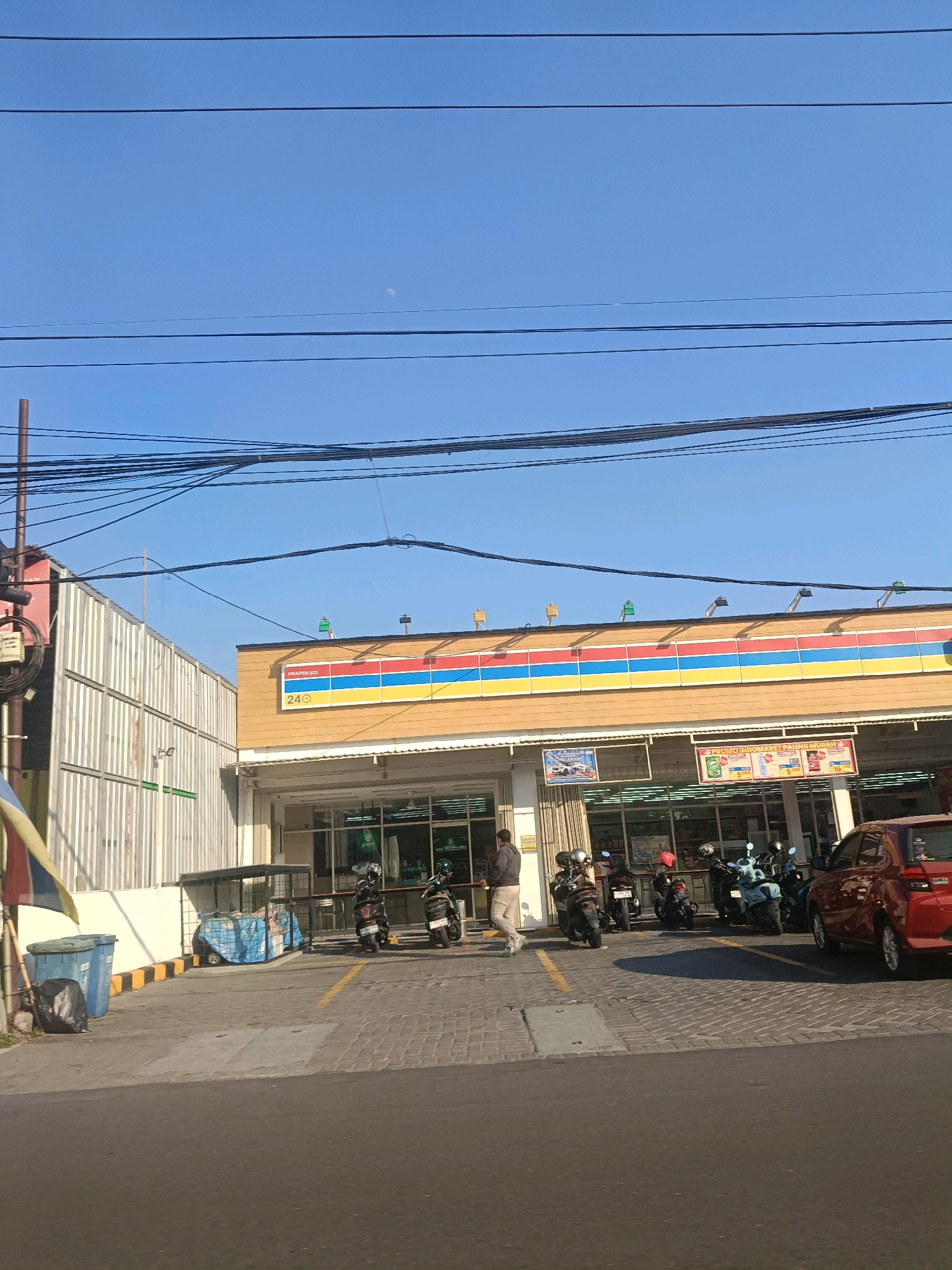 The image shows a street-level view of an Indomaret convenience store in Surabaya, Indonesia. The building has a distinctive yellow facade with red, blue, and yellow stripes across the top. A red car is parked in front of the store. Several motorcycles are also parked in the lot, and a few people are visible near the entrance. The sky is clear and blue with some power lines visible. The overall scene depicts a typical commercial area in a city.