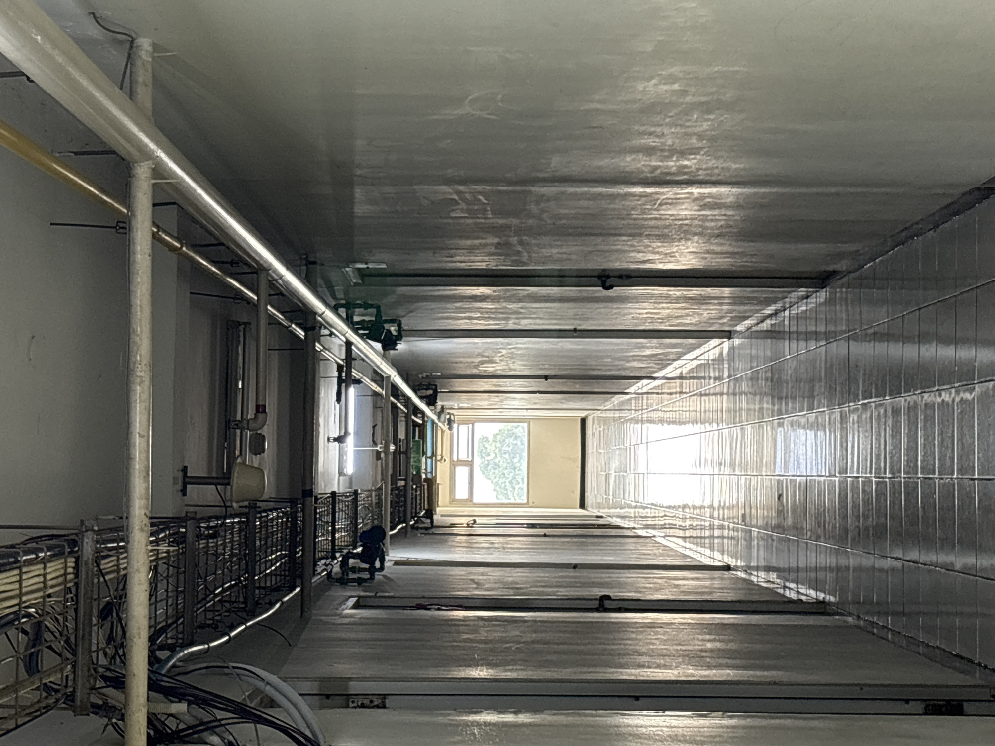 The image shows a long, narrow, dimly lit hallway with a tiled floor that reflects the overhead lighting. The walls are a light color and appear to be painted concrete. There are numerous pipes and conduits running along the ceiling and down the walls, suggesting an industrial or utility space. Several doors are visible on either side of the hallway. At the far end of the hallway, a window with a view of trees can be seen. The overall impression is one of a functional, perhaps older, building. The presence of the window at the end suggests this hallway might be located in a residential building or an office, as opposed to a purely industrial facility. Without further context, it is impossible to definitively place this specific hallway in North Jakarta, Indonesia.