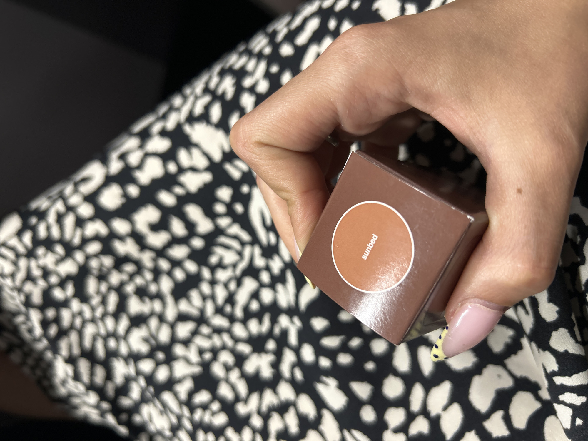 A close-up shot shows a person's hand holding a small, square, brown cosmetic box. The box has a circular window on its front, revealing a swatch of bronzer or blush in a warm, reddish-brown shade. Inside the circular window, the word "sunbed" is printed in white text. The person's fingernails are visible, with one nail adorned with a pale pink polish and yellow and black polka dots. The background consists of a black and white patterned fabric, possibly a dress or skirt, with an abstract, irregular spot design. The lighting appears to be indoor, with no strong shadows, suggesting it could be daytime. The overall impression is of a person showing off a beauty product.
