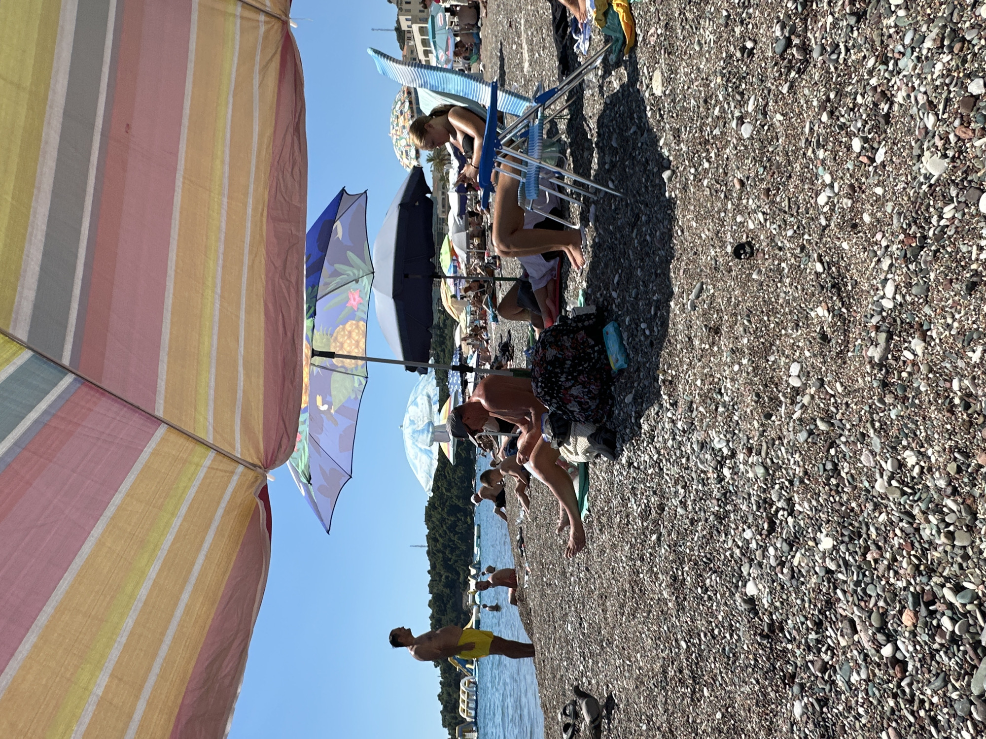 The image depicts a crowded beach scene on a sunny day in Boreti, Montenegro. The foreground is dominated by a pebbled shoreline. Several beach umbrellas provide shade for beachgoers. In the mid-ground, people are relaxing on beach chairs and towels. A woman in a blue striped chair is looking at her phone. A man in a dark shirt and hat is seated on the ground, also appearing to be engaged with his phone. Another man in yellow shorts stands by the water's edge, looking towards the sea. In the background, the clear blue water of the Adriatic Sea is visible, with inflatable water attractions in the distance. Trees and buildings line the coast. The overall atmosphere is one of leisure and summer recreation.