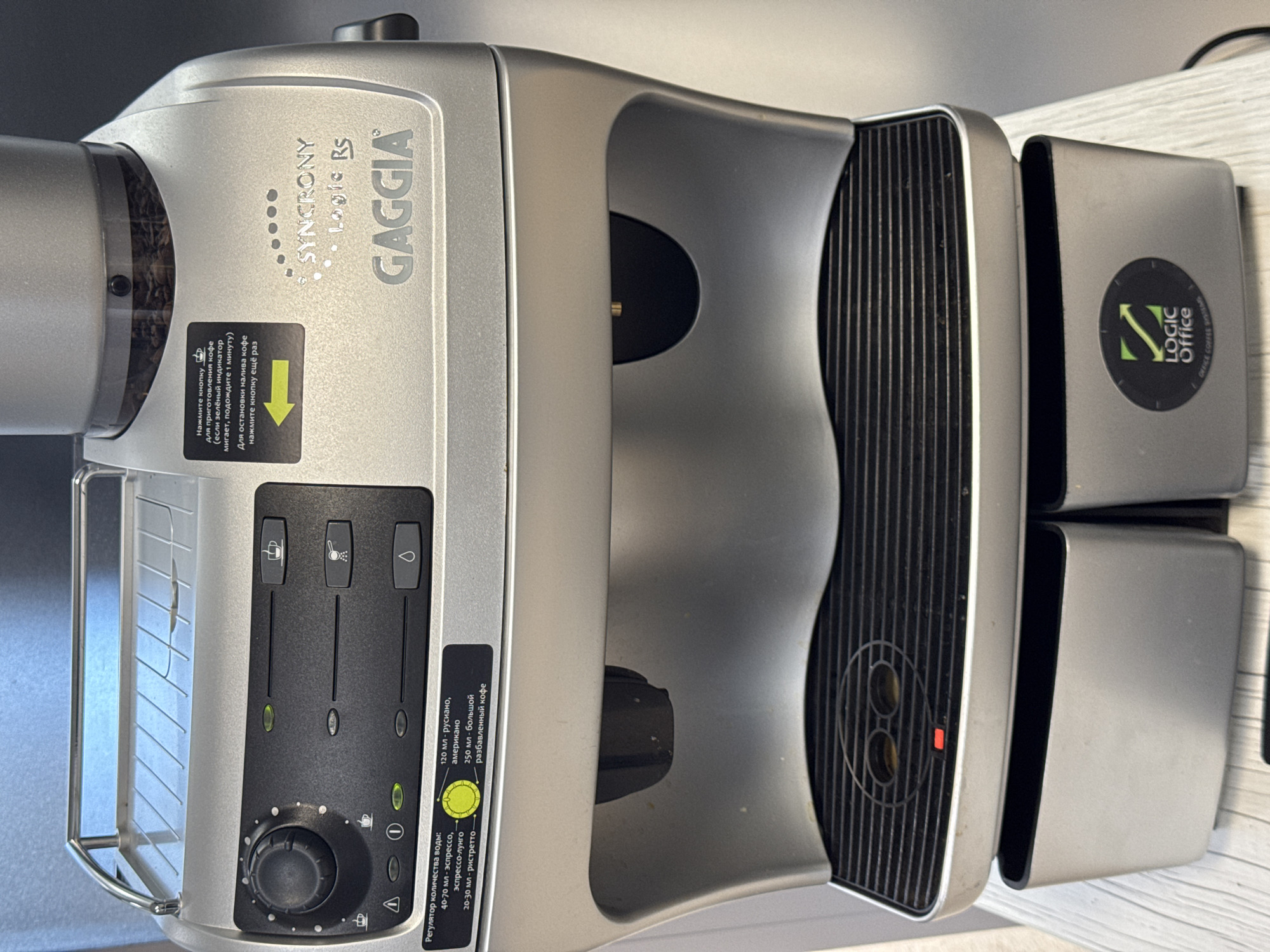 The primary subject of the image is a Gaggia Syncrony Logic RS automatic coffee machine, presented from an overhead perspective and tilted to its right. The machine is predominantly silver with black accents, featuring a modern and streamlined design. It rests on a light-colored, possibly wooden, countertop or table, likely indoors in Saint Petersburg, Russia.
The coffee machine's bean hopper, visible through a clear cover on the left, is filled with dark roasted coffee beans. Below this, a black instruction label in Russian provides operational guidance: "Press the button for coffee preparation (if the green indicator blinks, wait 1 minute)" and "To stop coffee pouring, press the button again."
The central control panel is black, featuring three vertical buttons with distinct pictograms for different functions (e.g., coffee cup, steam/hot water, water drop), each accompanied by a green indicator light. Below these is a rotary dial, potentially for adjusting coffee strength or grind, marked with a small warning triangle. A yellow sticker adjacent to the dial specifies water volume settings in Russian: "Water quantity regulator: 40-70 ml - espresso, espresso-lungo; 20-30 ml - ristretto; 120 ml - Americano; 250 ml - large diluted coffee."
On the far right side of the machine, a circular black sticker with a green "Z LOGIC Office" logo is visible, suggesting a specific product line or intended use in an office environment. The coffee dispensing area includes a black grate with two spouts and a red indicator light. The machine appears clean and well-maintained, ready for brewing coffee.